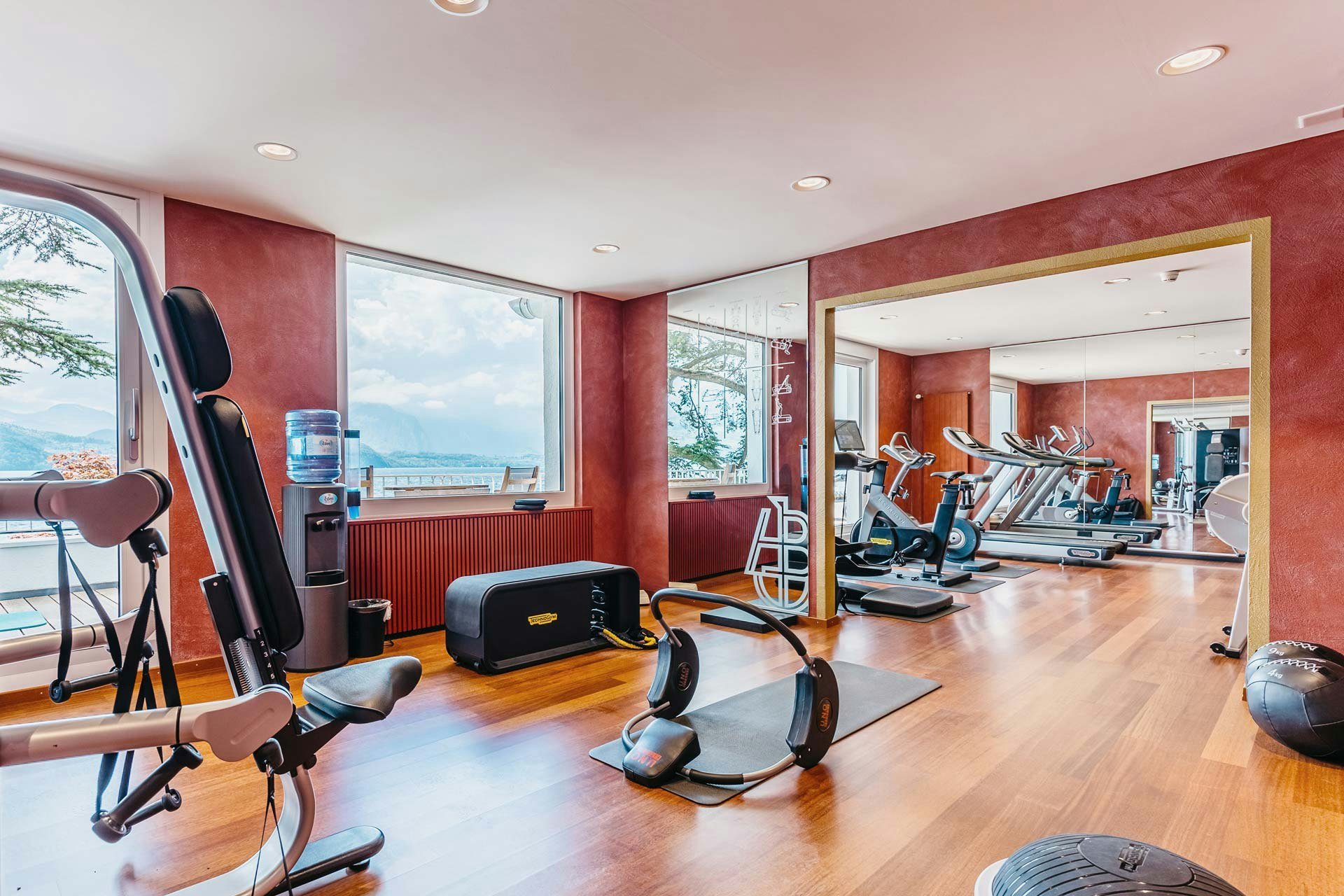 Salle avec sol en bois brillant, grand mur de miroirs et fenêtre panoramique avec divers appareils de fitness