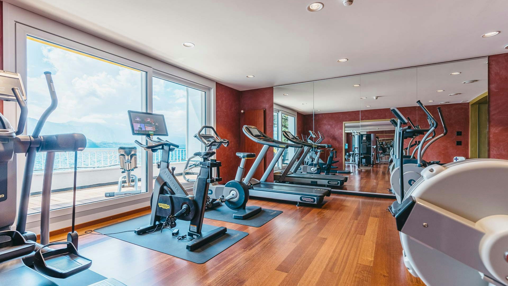 Fitness-Studio im Hotel BEATUS Raum mit glänzendem Holzboden, grosser Spiegelwand und Panoramafenster mit diversen Fitnessgeräten