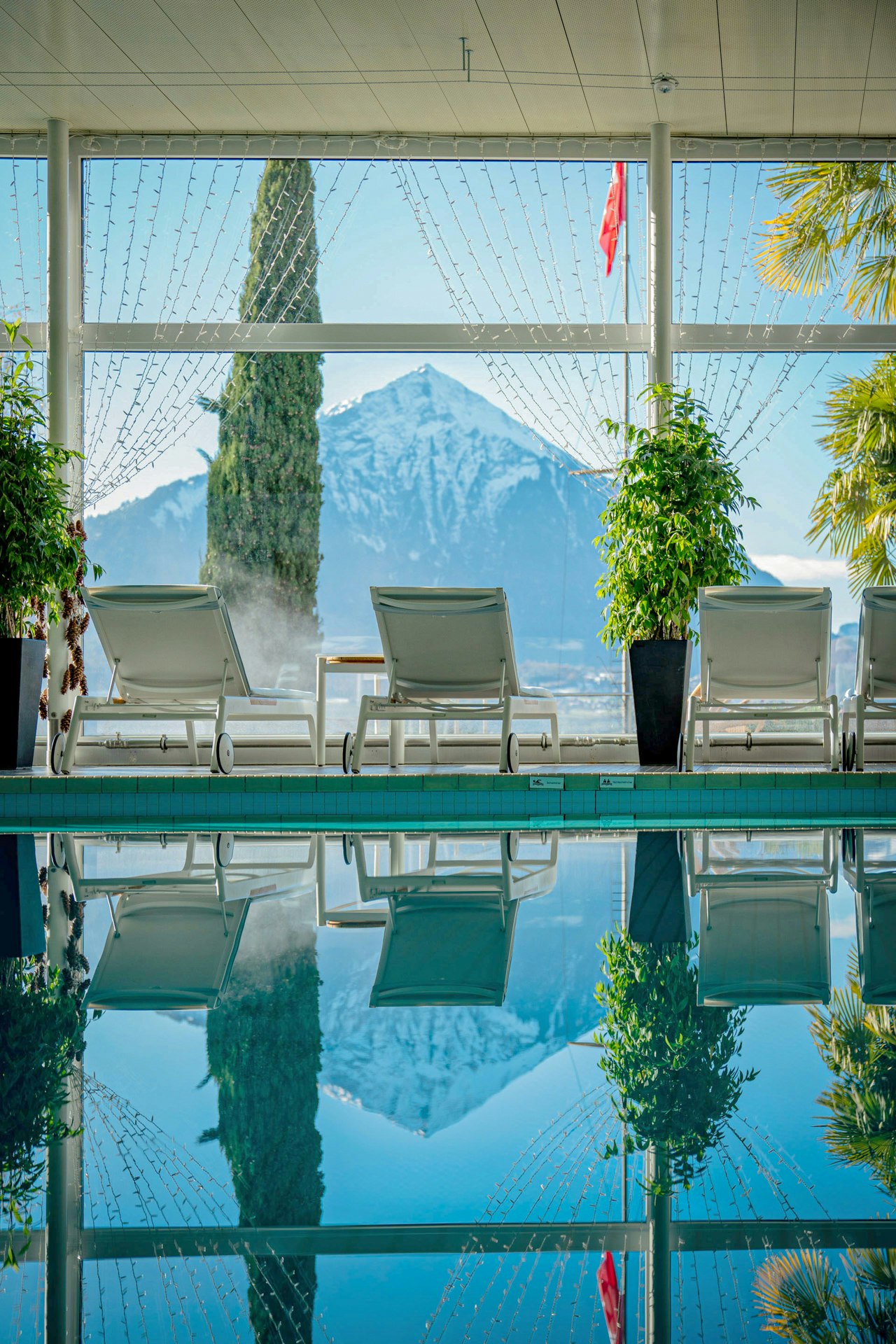 Vorne das blaue Wasser des Hallenbades, darin spiegelt sich der Berg, den man auch durch das Fenster sieht, dazwischen Liegestühle