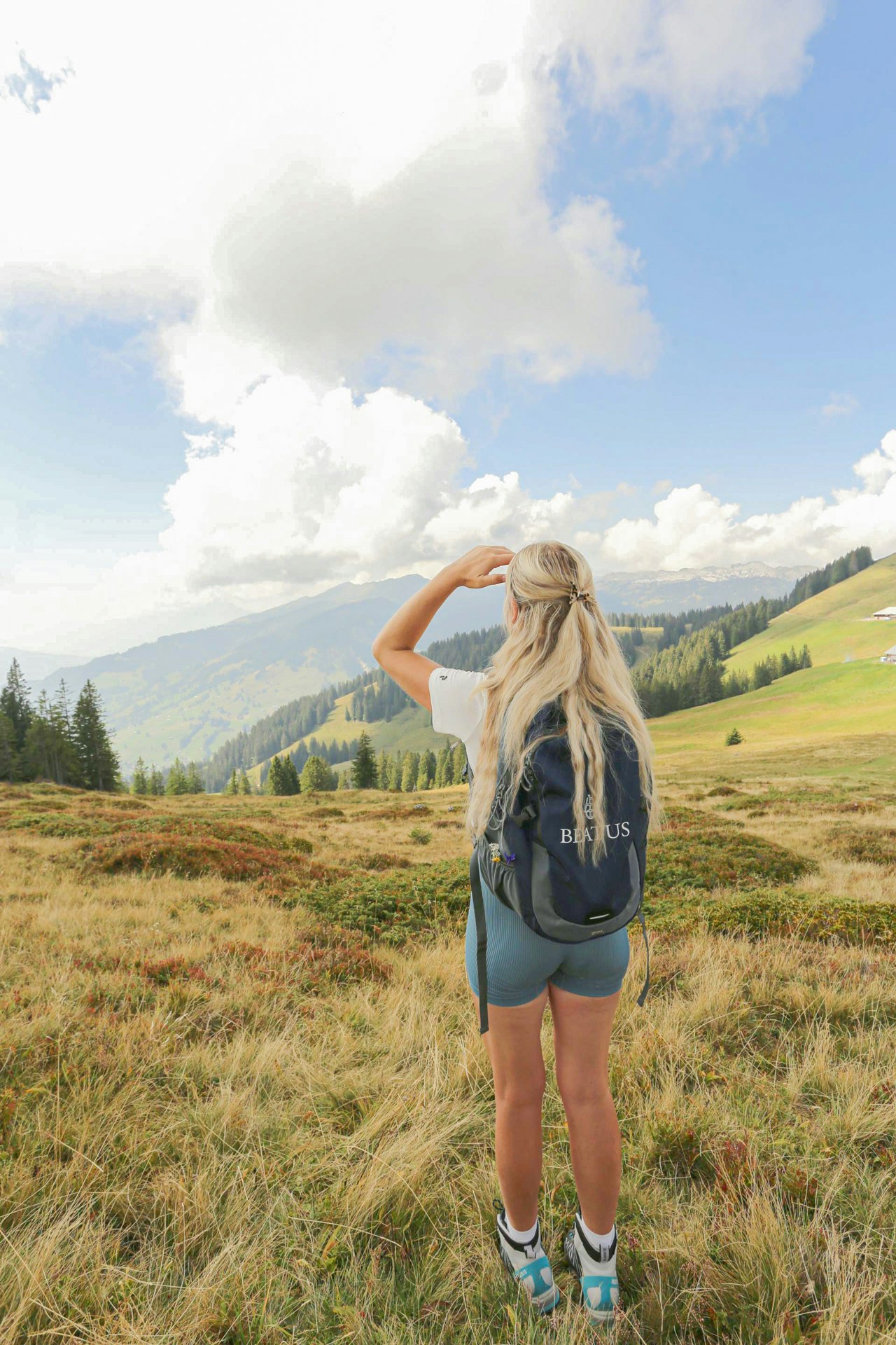 Eine Frau mit kurzen Hosen, Wanderschuhen, Rucksack und langen blonden Haaren blickt über eine Alpwiese ins Tal