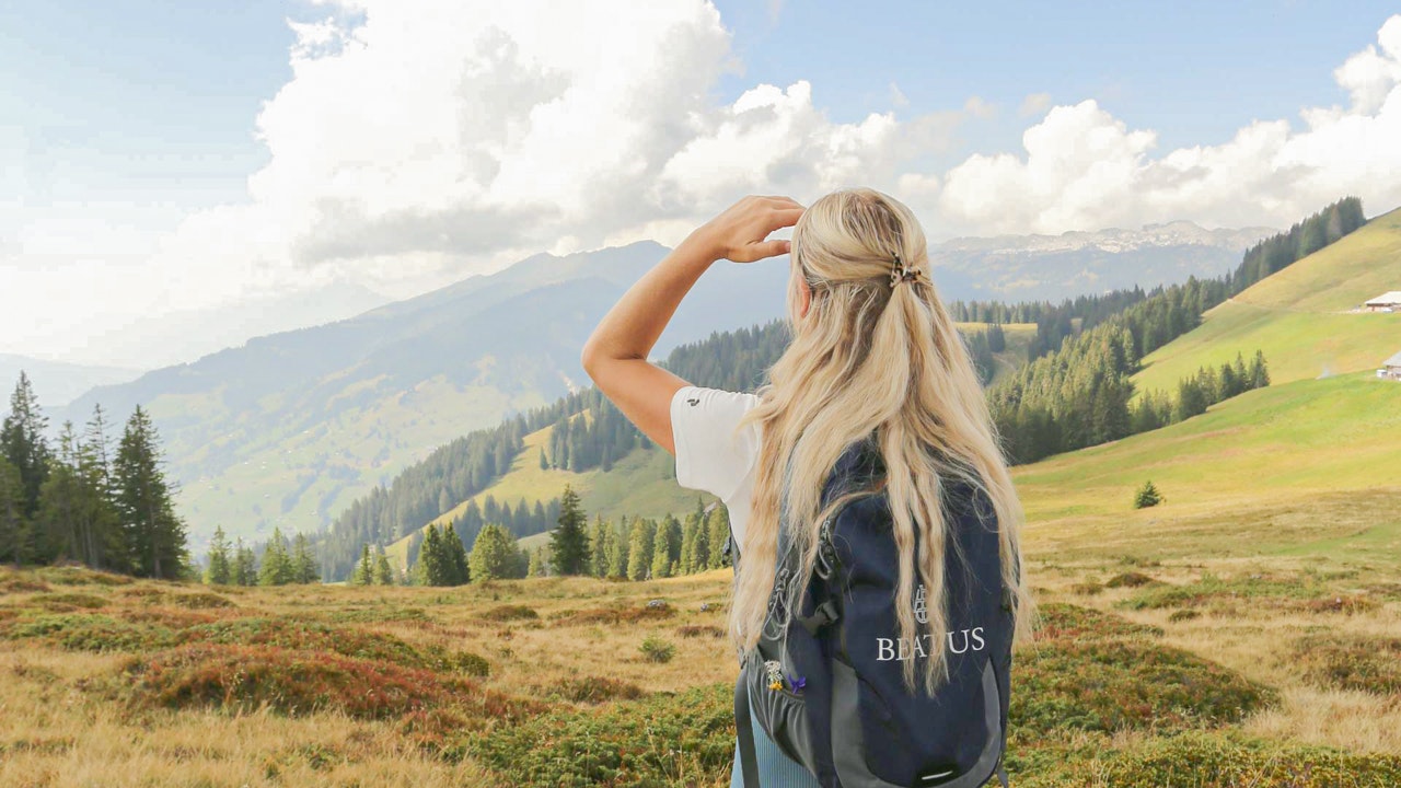 Unterwegs mit den Natur-Guides Eine Frau mit kurzen Hosen, Wanderschuhen, Rucksack und langen blonden Haaren blickt über eine Alpwiese ins Tal