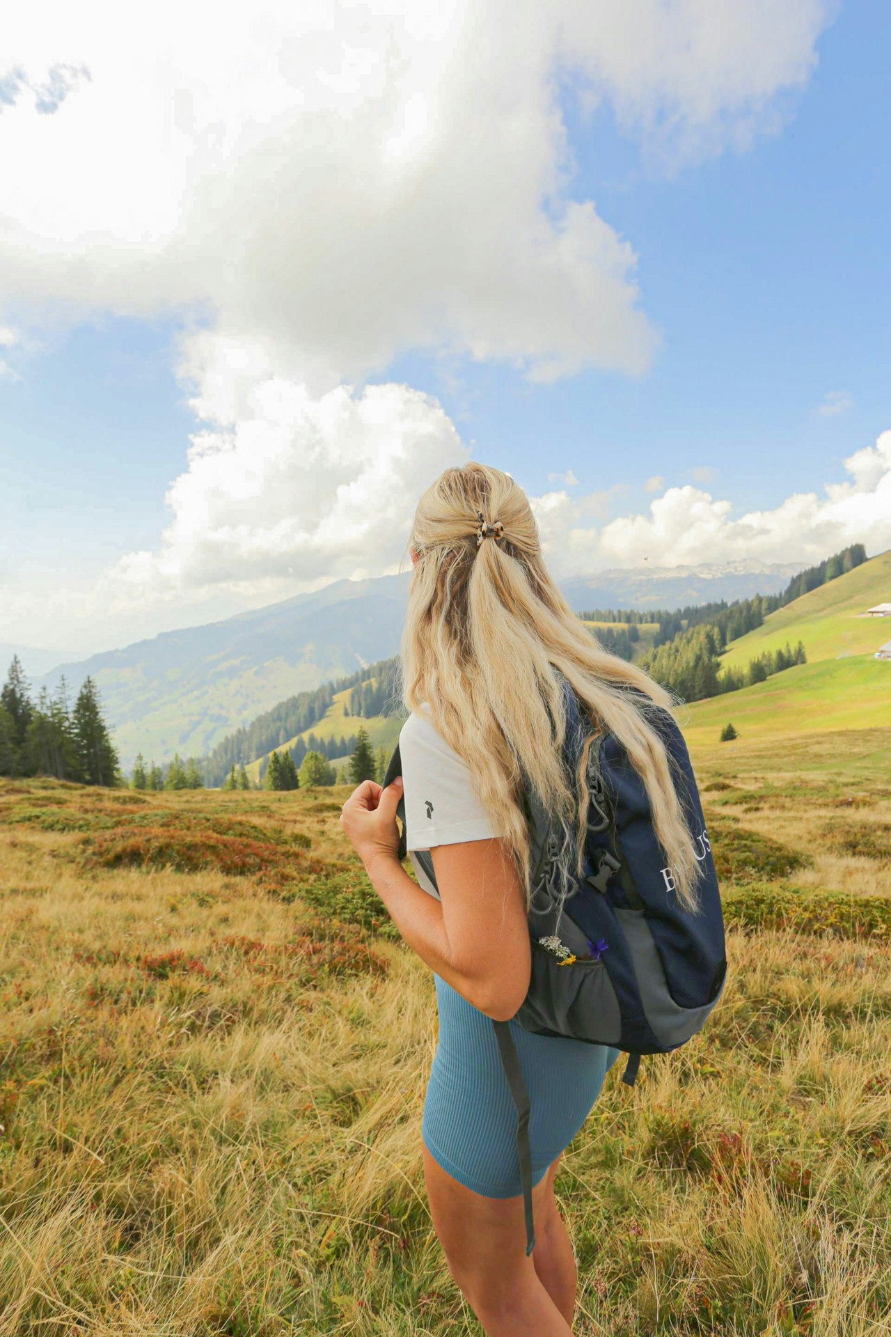 A woman with a rucksack and shorts looks out over the valley from a mountain meadow