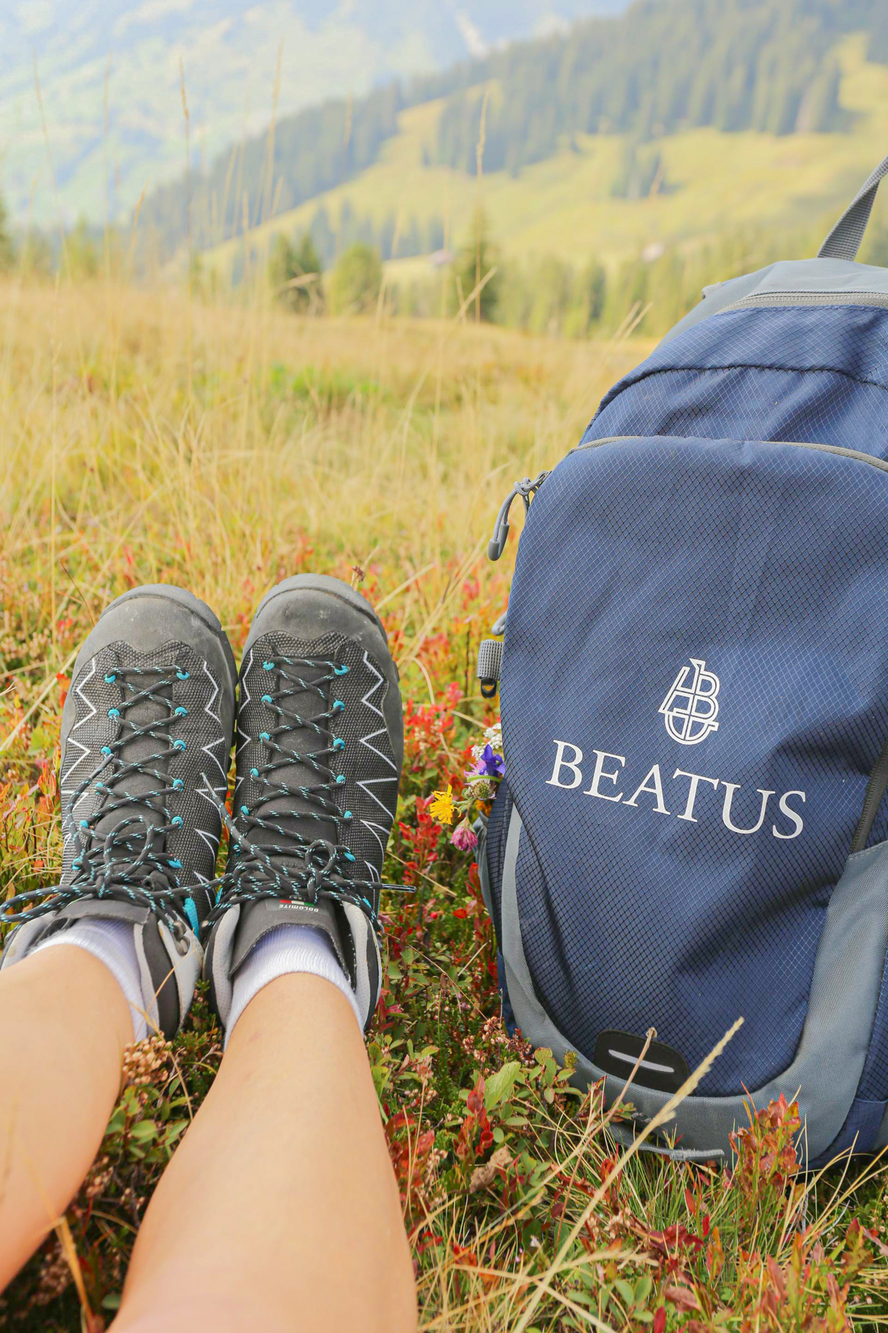 Jemand sitzt in einer Bergwiese, man sieht nur die Wanderschuhe, rechts ein Rucksack mit der Aufschrift Beatus