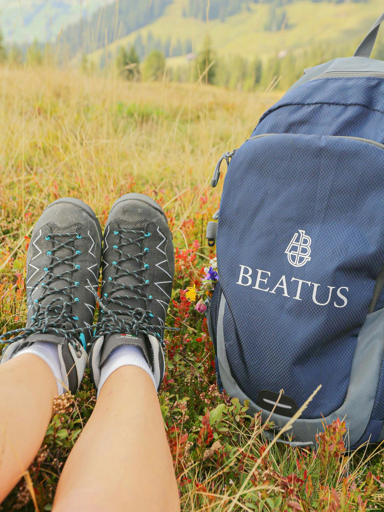 Unterwegs mit den Natur-Guides Jemand sitzt in einer Bergwiese, man sieht nur die Wanderschuhe, rechts ein Rucksack mit der Aufschrift Beatus