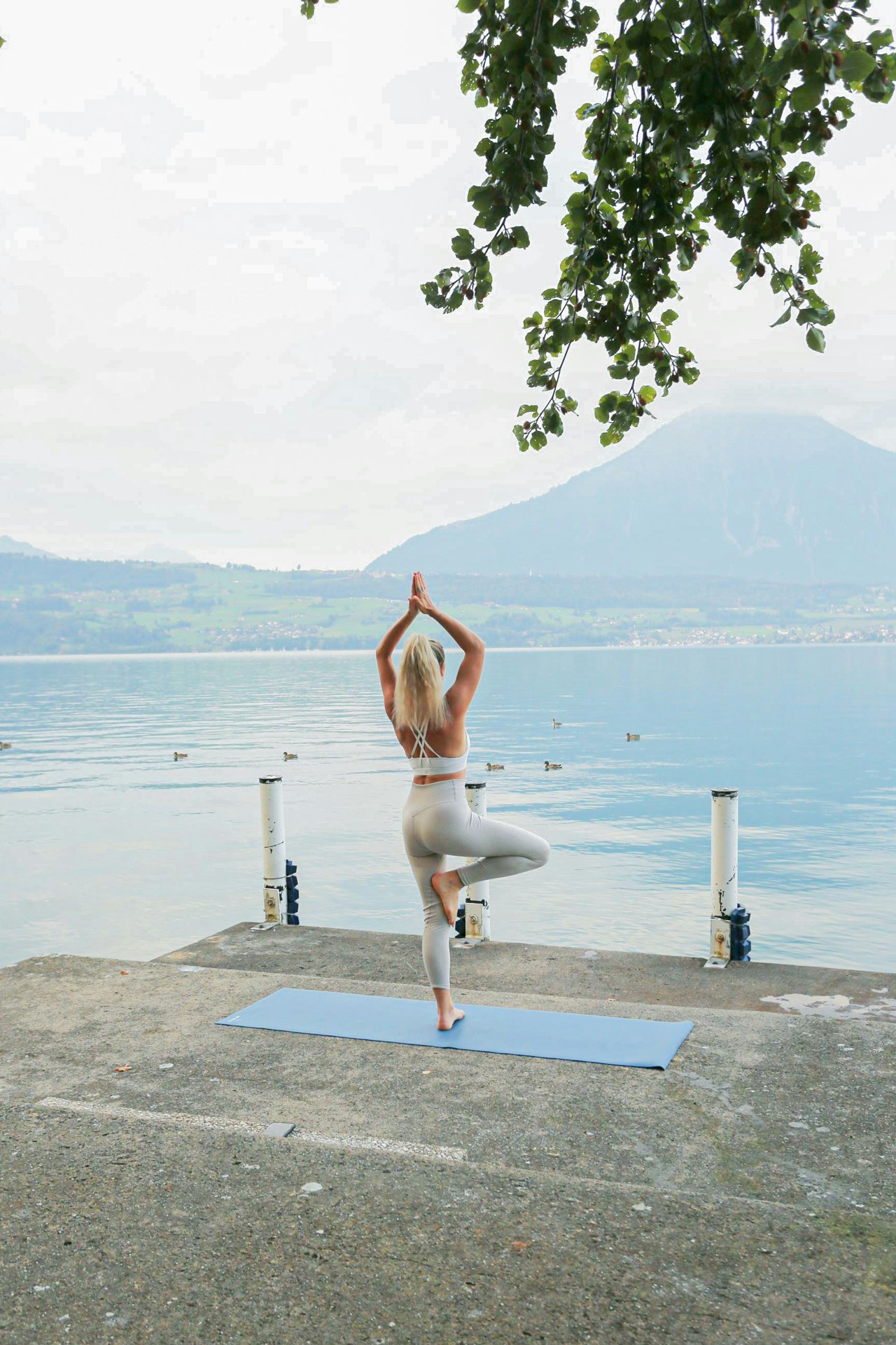 Eine Frau steht in einer Yogaposition auf einer blauen Yogamatte vor dem Thunersee