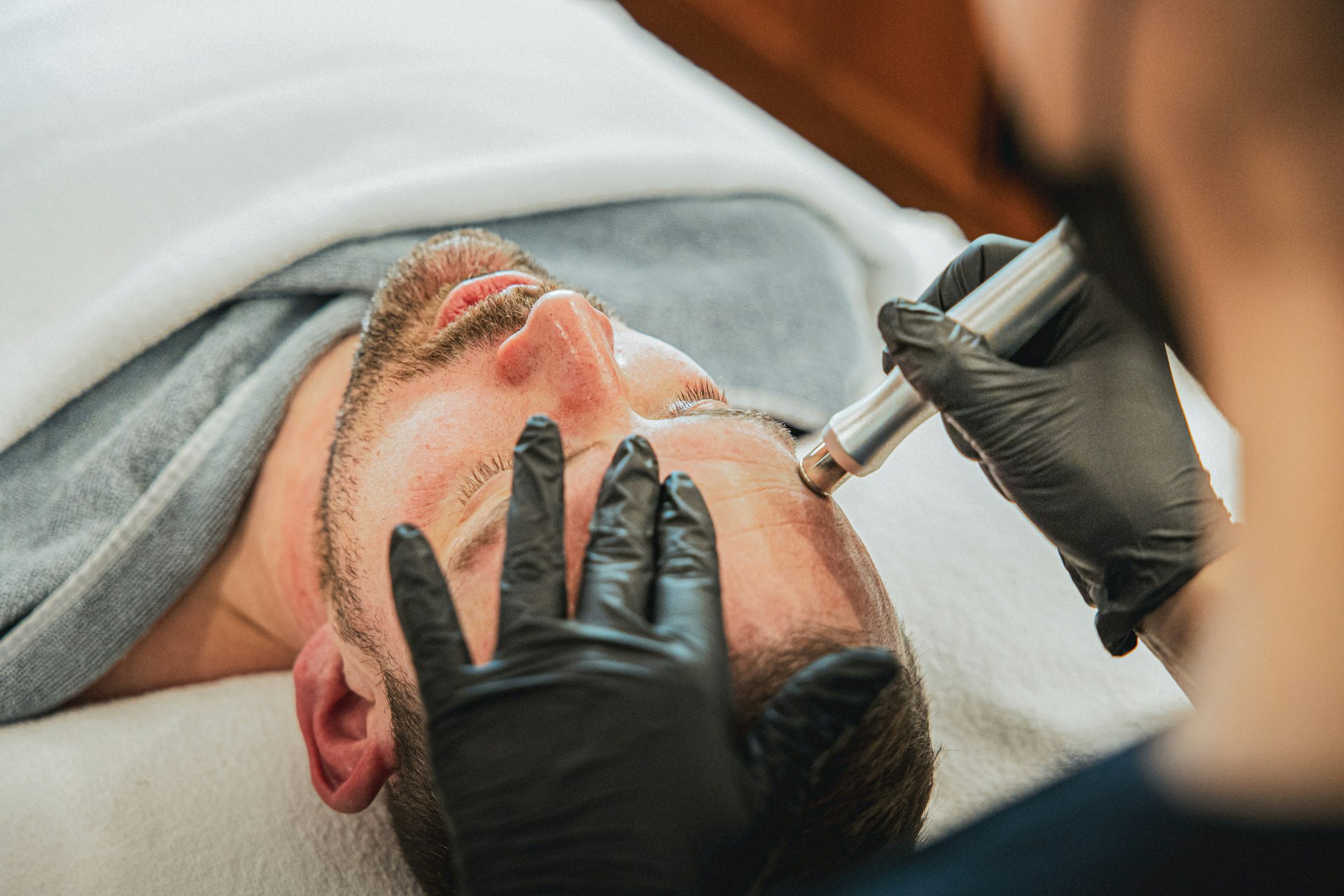 Ein Mann liegt auf einer Massageliege, bedeckt mit Badetüchern, sein Gesicht wird von einem anderen Mann hinter ihm mit einem Metallstift behandelt