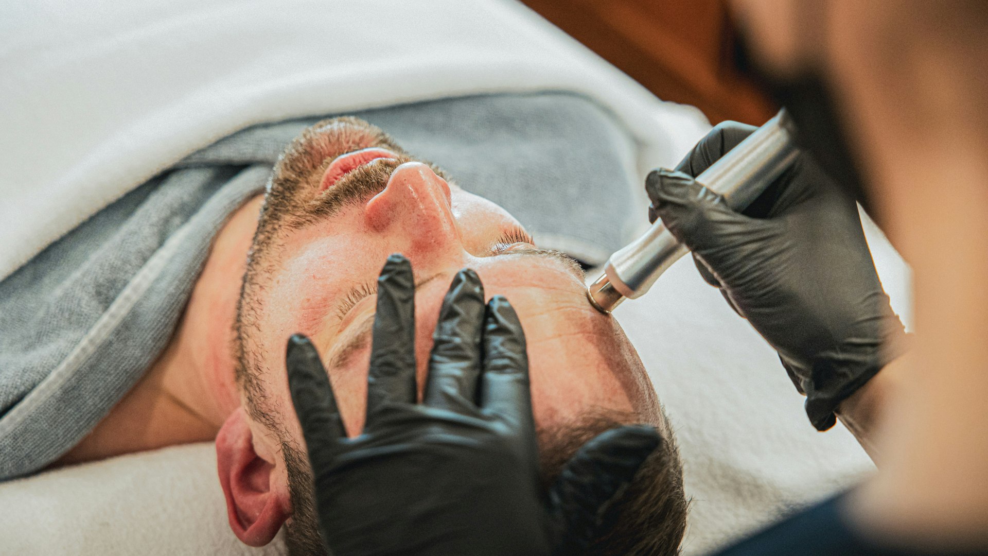 Beautybehandlungen für den Mann im Hotel BEATUS Ein Mann liegt auf einer Massageliege, bedeckt mit Badetüchern, sein Gesicht wird von einem anderen Mann hinter ihm mit einem Metallstift behandelt