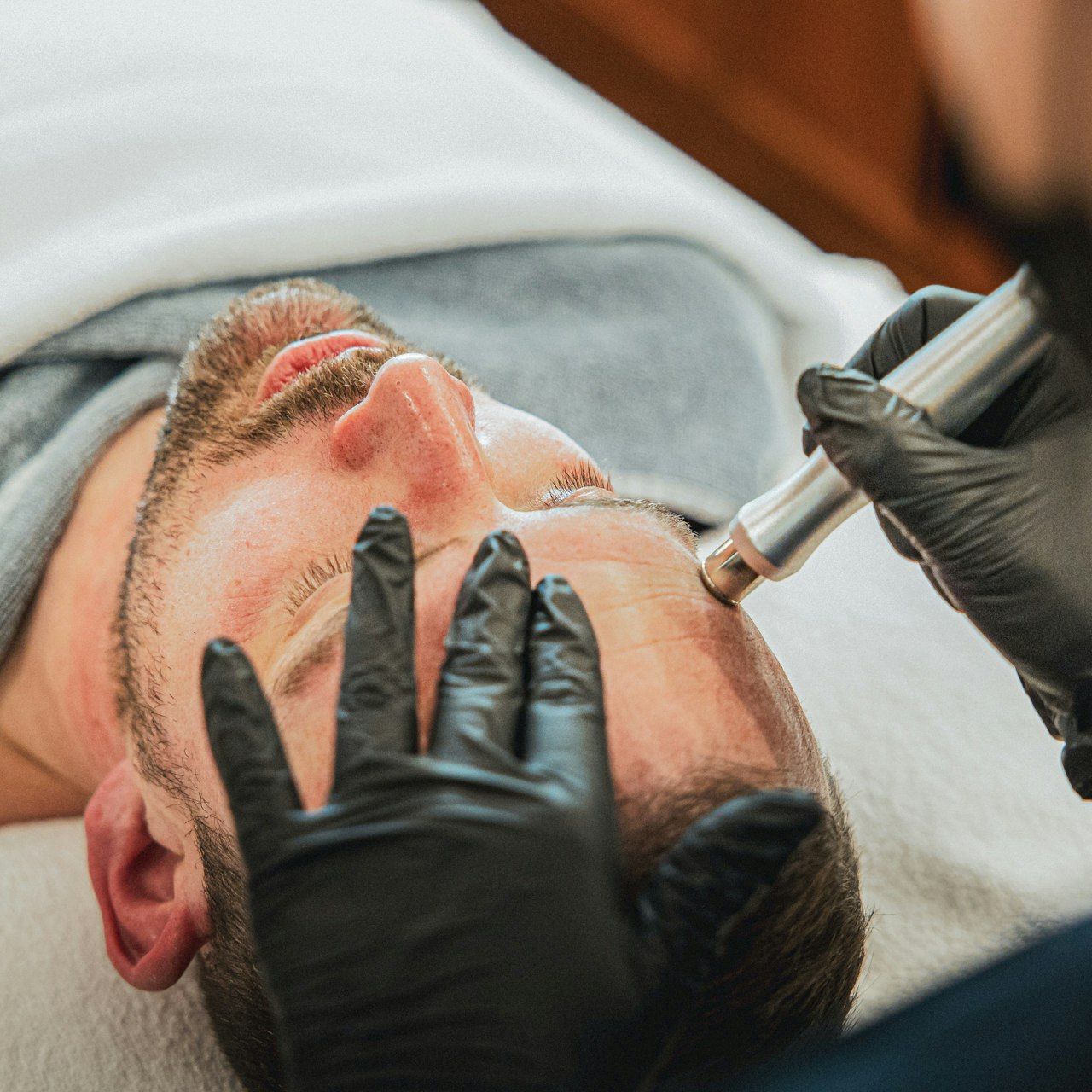 Beautybehandlungen für den Mann im Hotel BEATUS Ein Mann liegt auf einer Massageliege, bedeckt mit Badetüchern, sein Gesicht wird von einem anderen Mann hinter ihm mit einem Metallstift behandelt