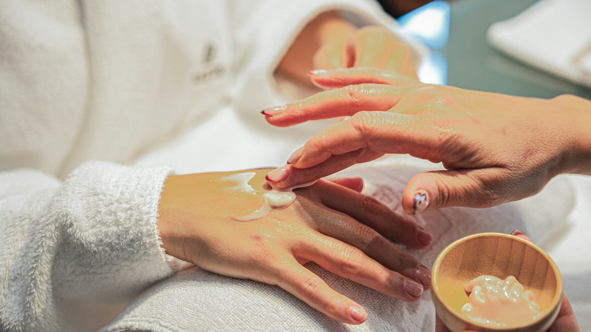 Maniküre im Hotel BEATUS Eine Frau im Bademantel hat ihre Hände auf einer Rolle abgelegt, eine andere Frau streicht sie mit einer Creme ein