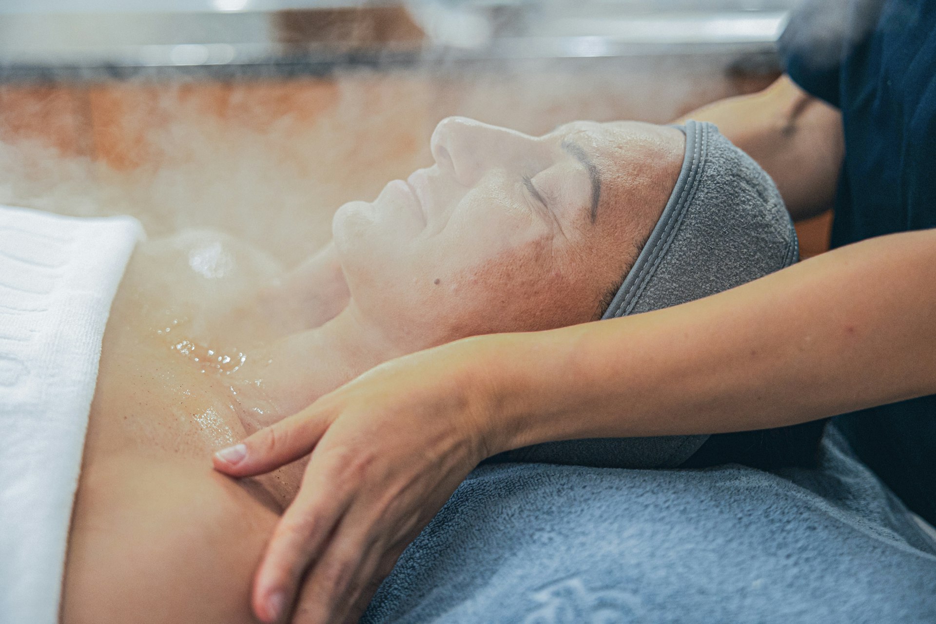 Eine Frau liegt auf einer Massageliege, ihr Gesicht im Dampf, dahinter eine Masseurin, die ihre Schultern massiert