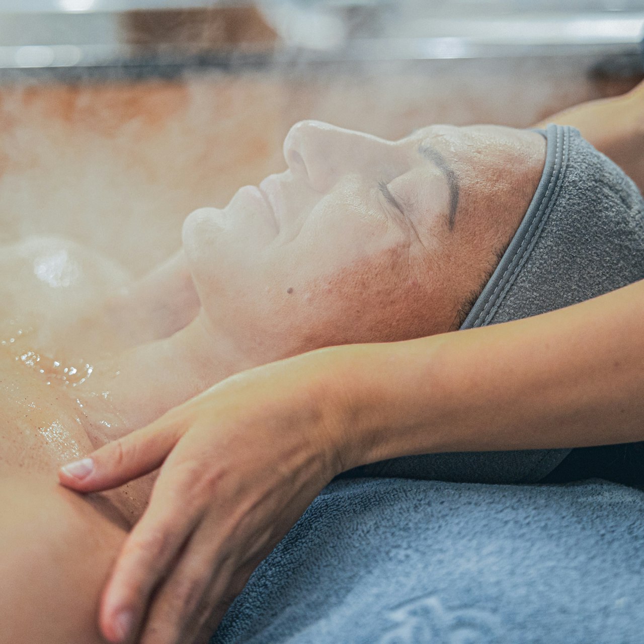 Beautybehandlungen im Hotel BEATUS Eine Frau liegt auf einer Massageliege, ihr Gesicht im Dampf, dahinter eine Masseurin, die ihre Schultern massiert