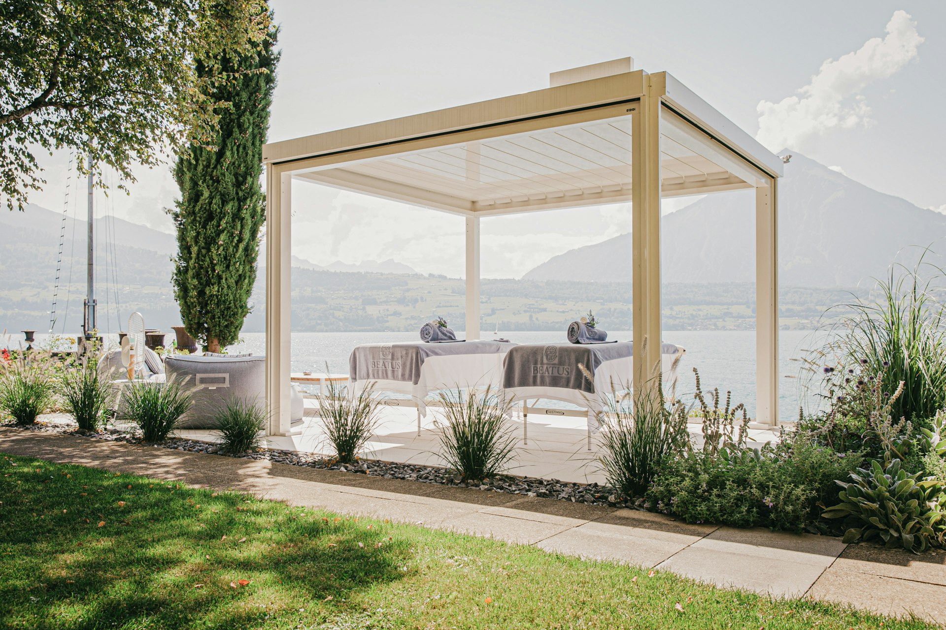 Ein überdachter Platz in der Sonne, darunter zwei Massageliegen mit Badetüchern, dahinter der See und die Berge