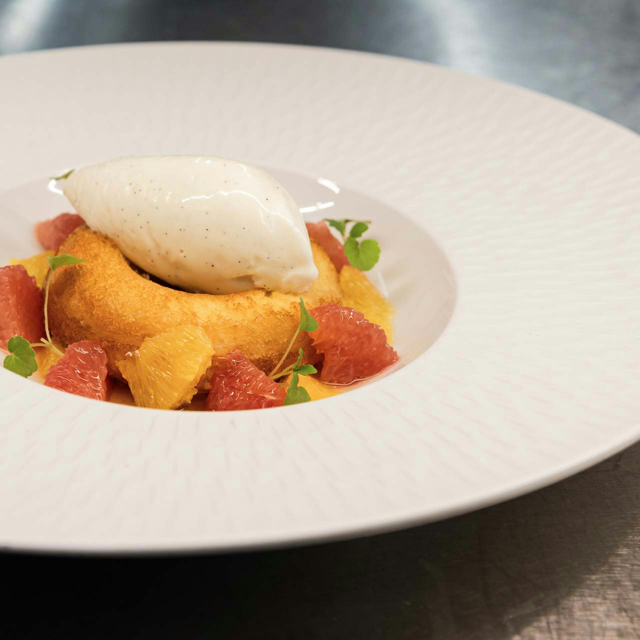 BEATUS Kulinarik Abendessen Weisser Teller auf Metalluntergrund, darin Orangenfilets, Teigring und Vanilleeis