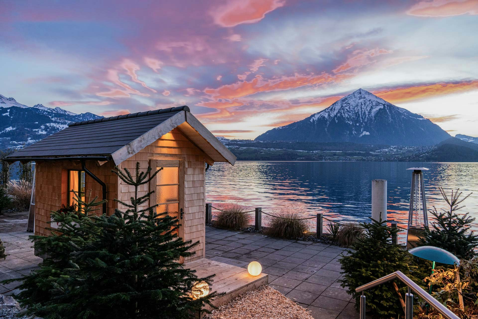 Wooden hut with light, two conifers in front of it, Lake Thun and Niesen in the twilight behind it