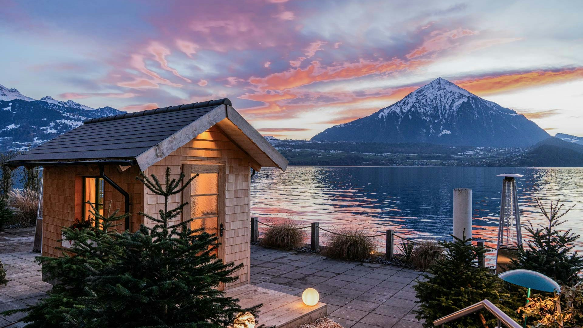 Fonduehüsli in der Dämmerung Holzhäuschen mit Licht, davor zwei Nadelbäume, dahinter der Thunersee und Niesen in der Dämmerung