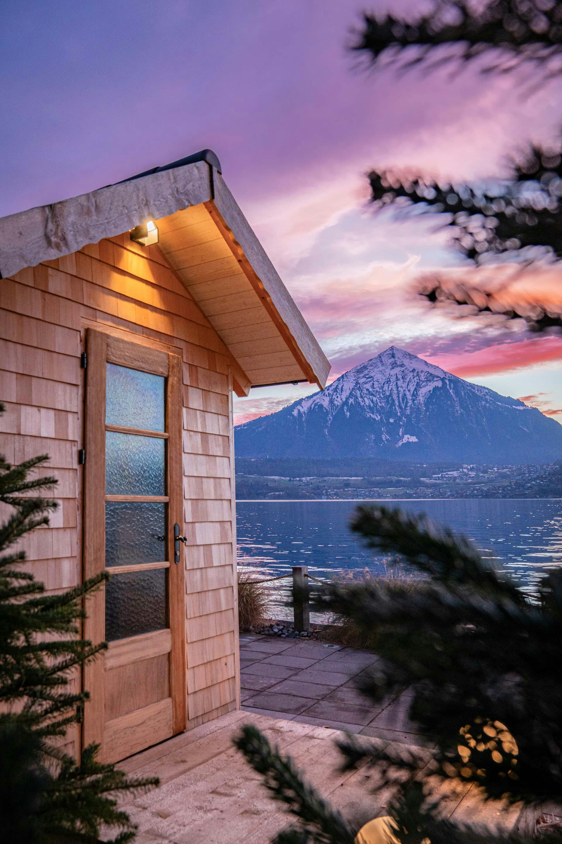 Holzhäuschen mit Licht, davor zwei Nadelbäume, dahinter der Thunersee und Niesen in der Dämmerung