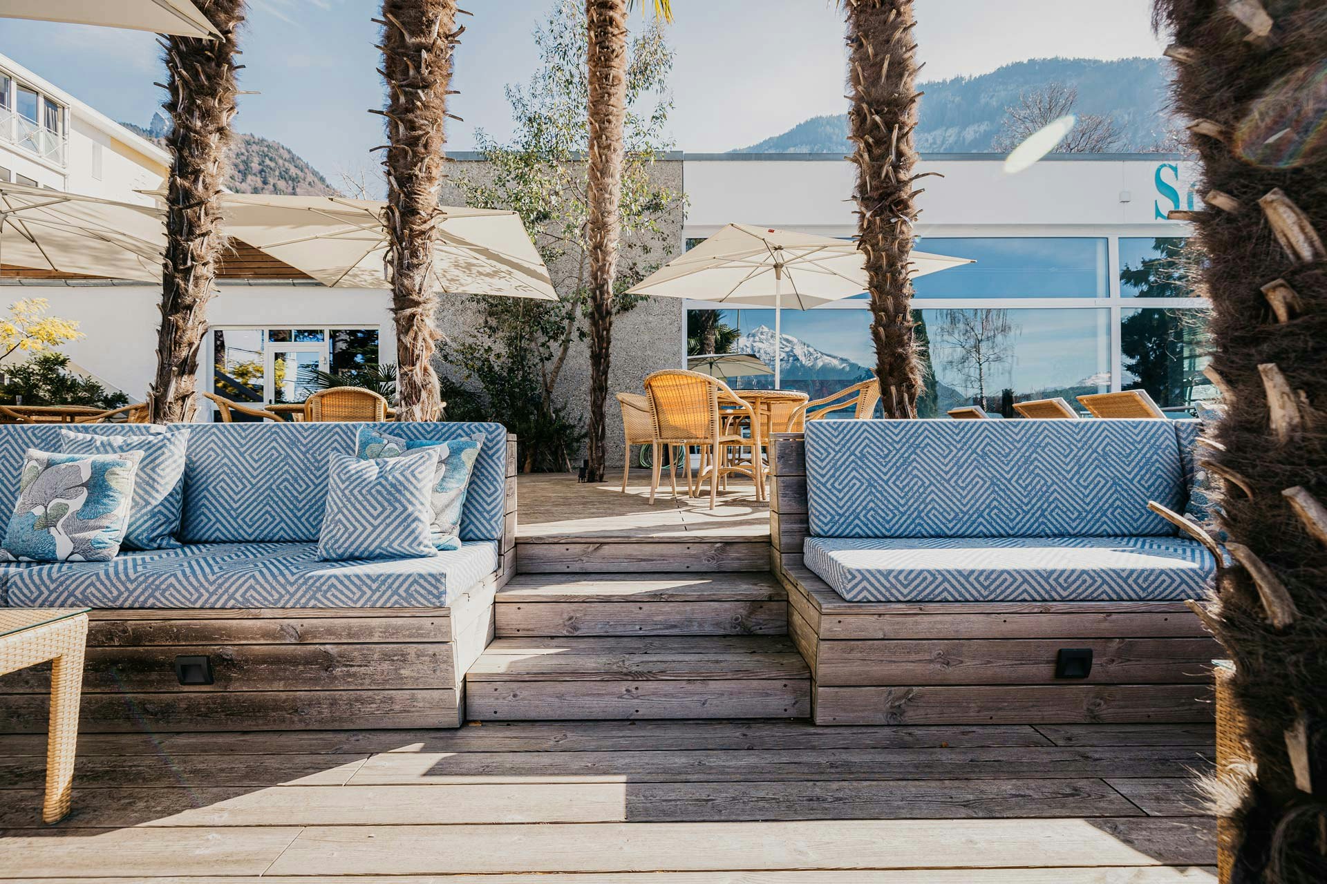 Sièges bleus et blancs, terrasse en bois, palmiers, chaises tressées à l'arrière, tables en verre, parasols et un bâtiment entaillé