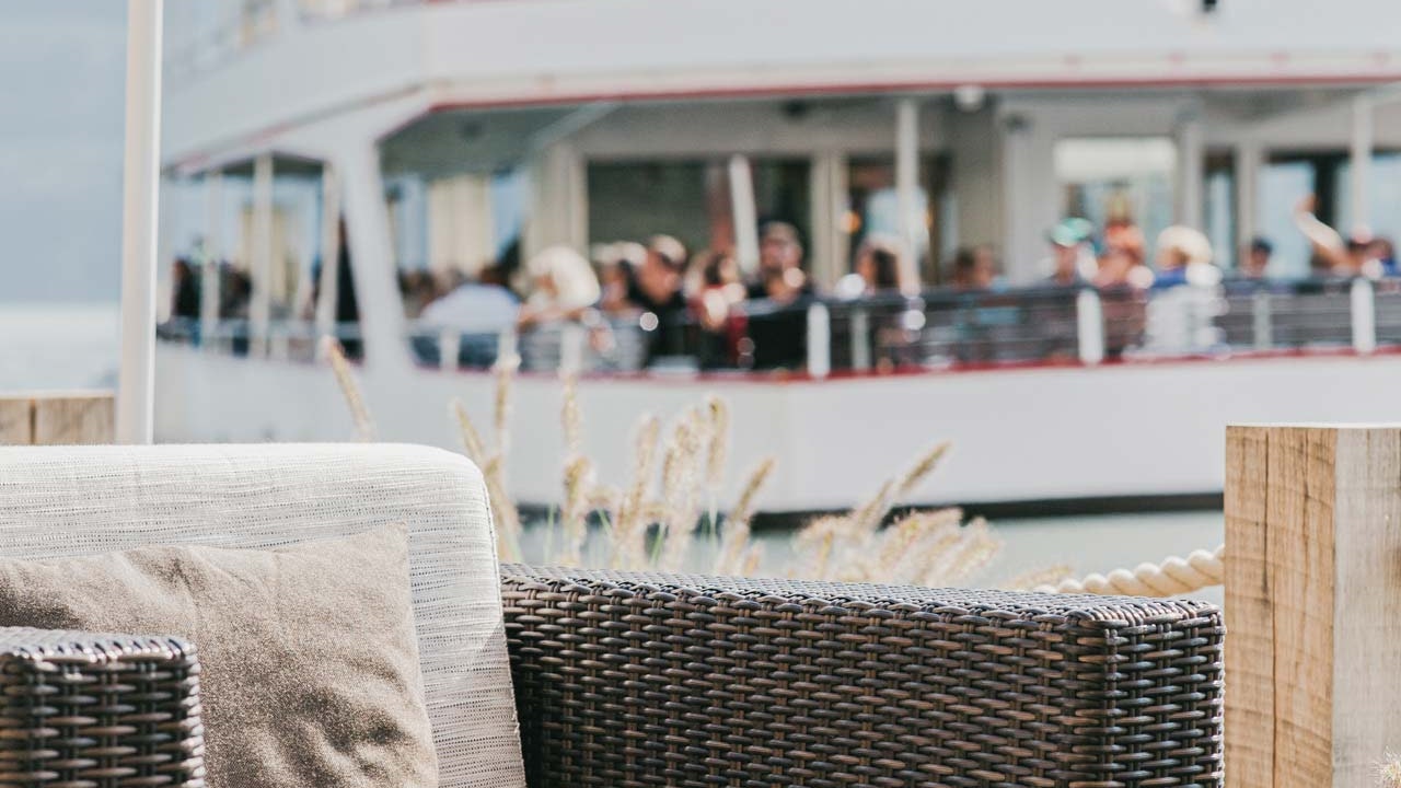 SeeLounge im Hotel BEATUS Loungemöbel und Stehlampe, dahinter in der Unschärfe ein Ausflugsschiff mit Menschen von Nahem