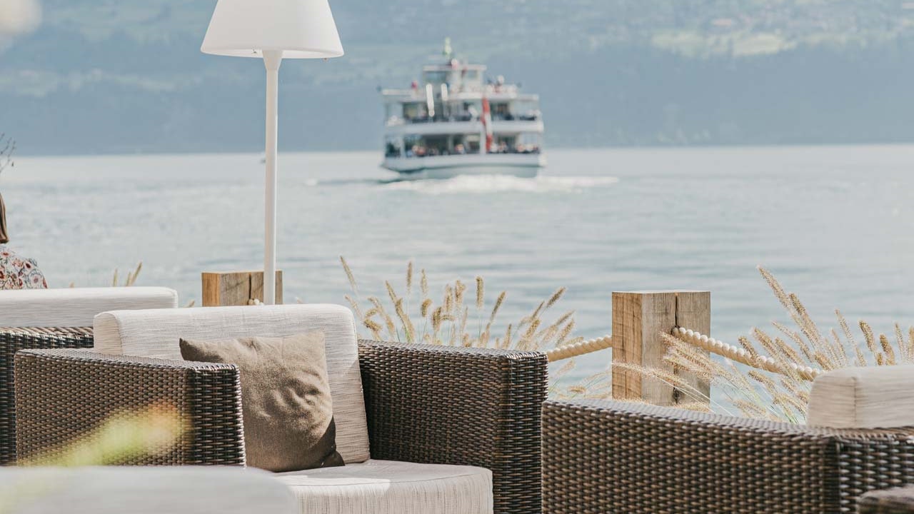 SeeLounge im Hotel BEATUS Geflochtene Loungmöbel, Sonnenschirme und weisse Stehlampe, dahinter der See mit einem Ausflugsschiff und ein Berg