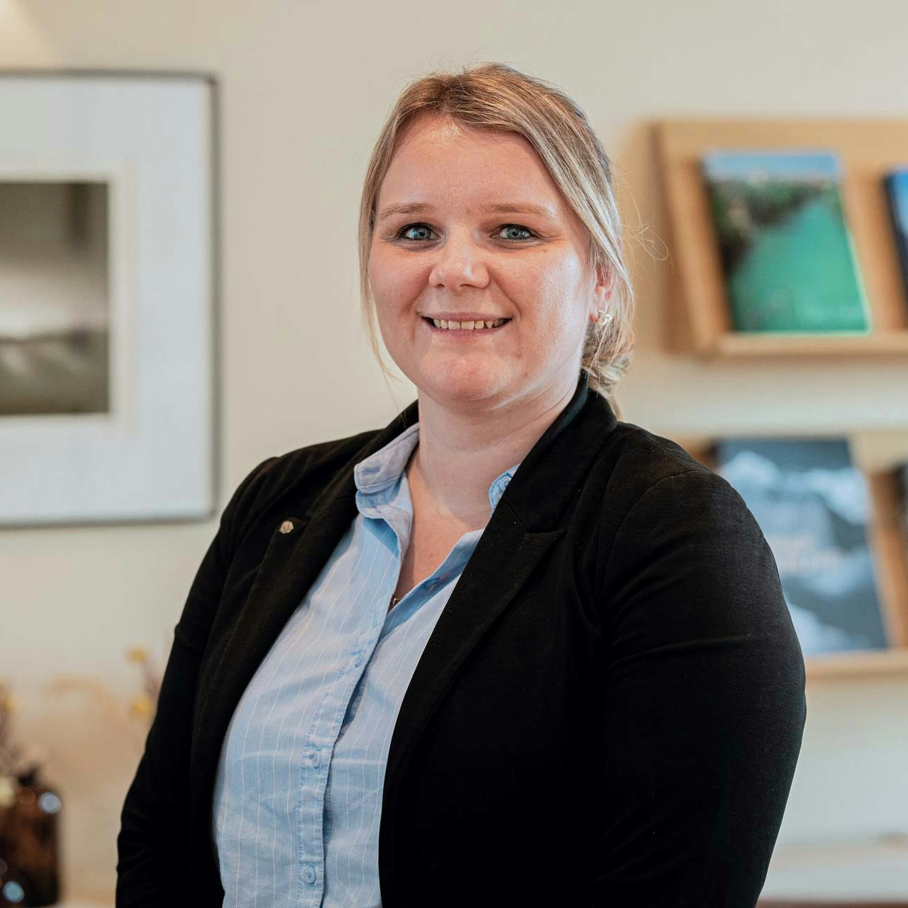 Sibylle Michel, Chef de Réception Frau mit blauer Bluse und schwarzer Weste vor Regal mit Magazinen