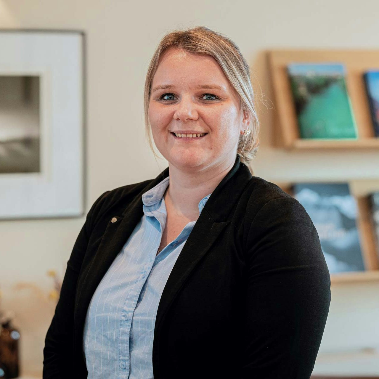 Sibylle Michel, chef de la réception Femme en chemisier bleu et gilet noir devant une étagère de magazines