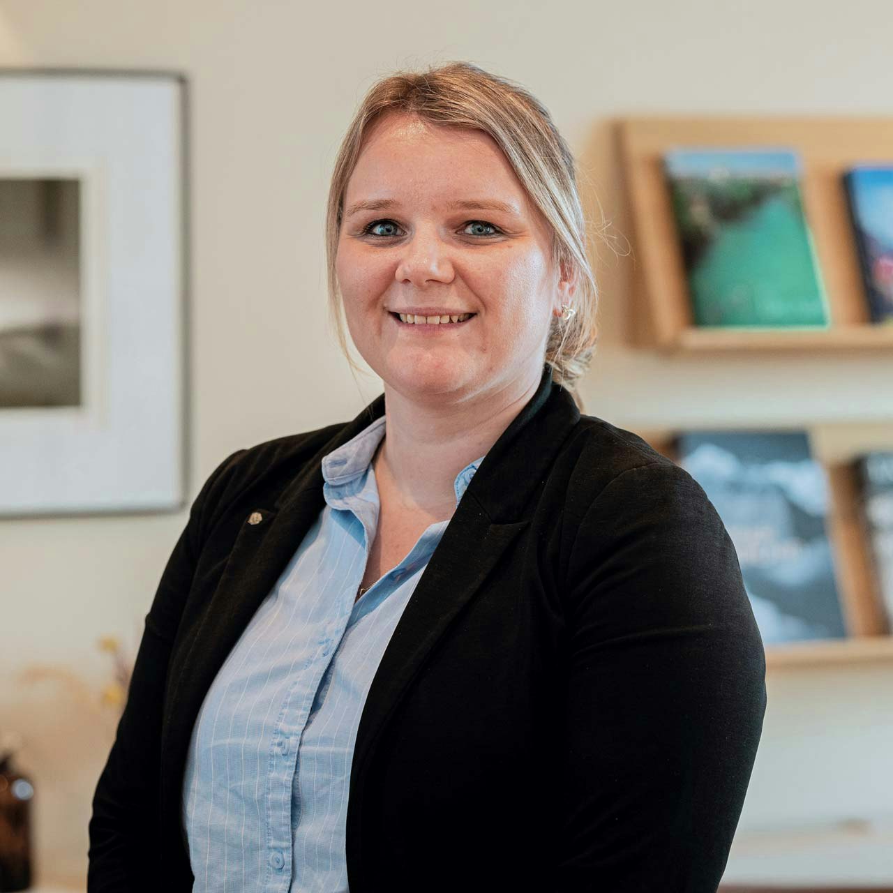 Sibylle Michel, Chef de Réception Woman with blue blouse and black waistcoat in front of a shelf with magazines