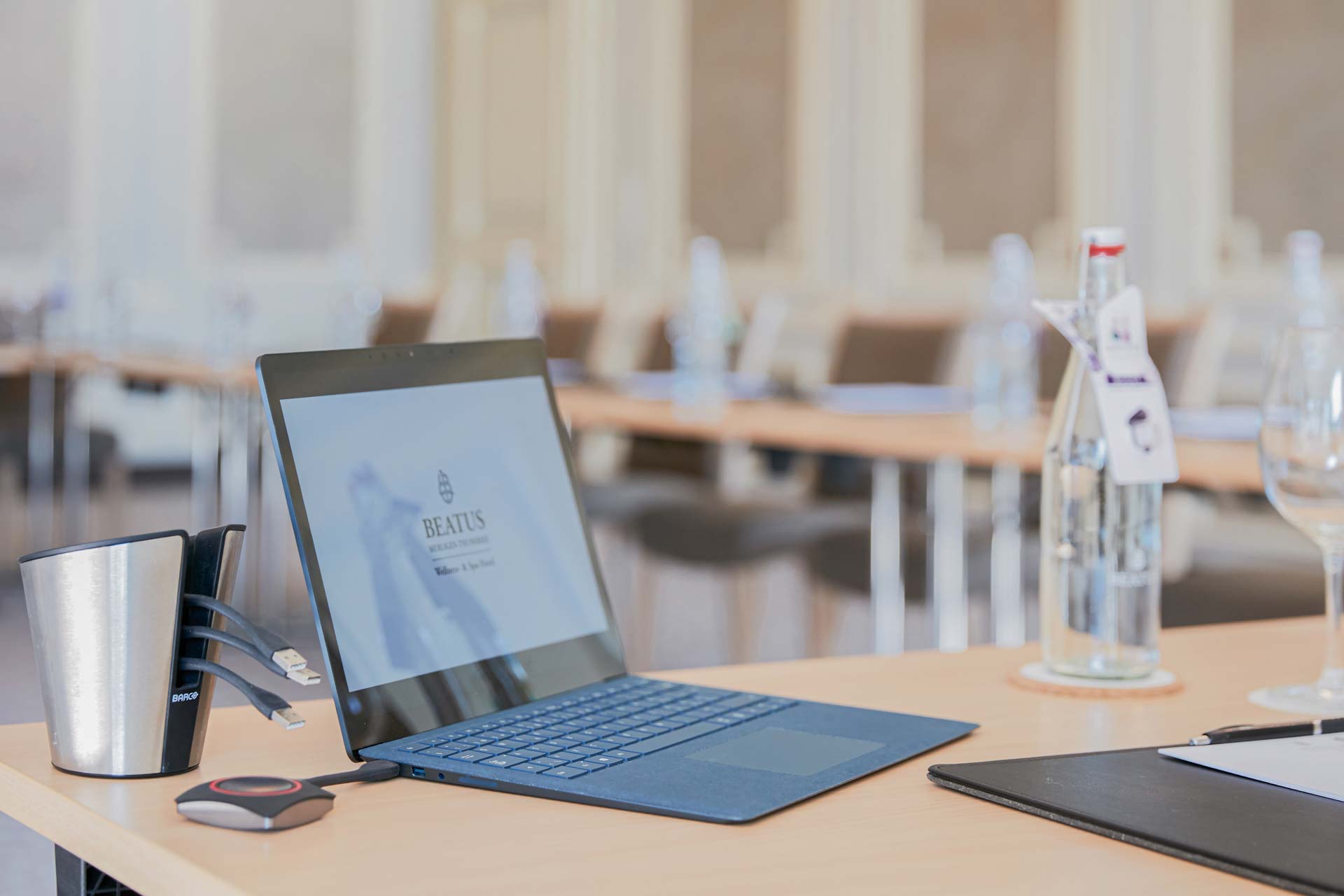 Ordinateur portable ouvert et différentes prises sur une table, à droite et au fond de l'image d'autres tables, bouteille en verre
