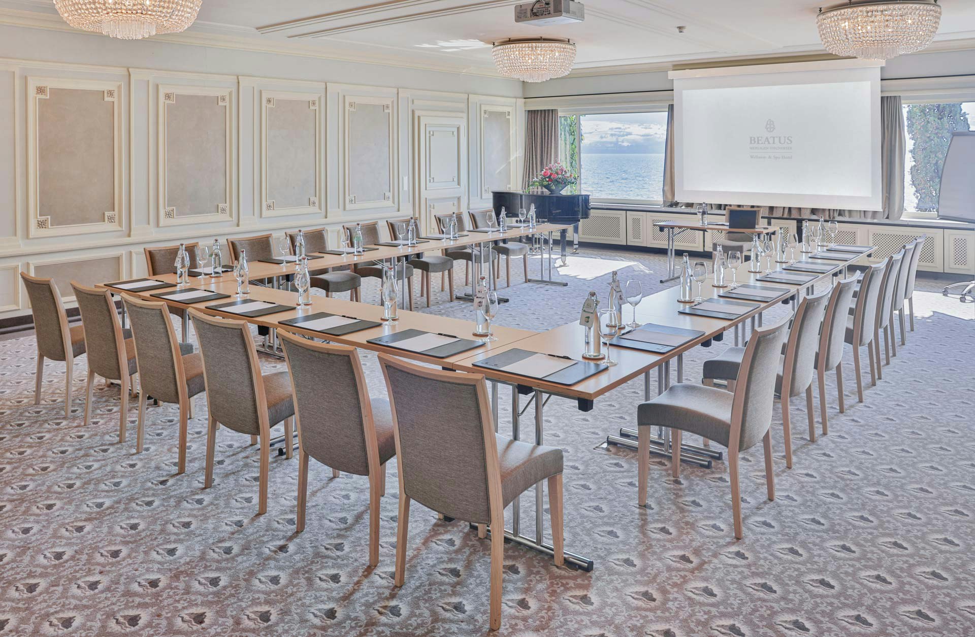 Grande salle avec moquette à motifs, motifs ronds en cristal, boiseries blanches et grandes fenêtres panoramiques, tables et chaises au centre, flipchart à l'avant