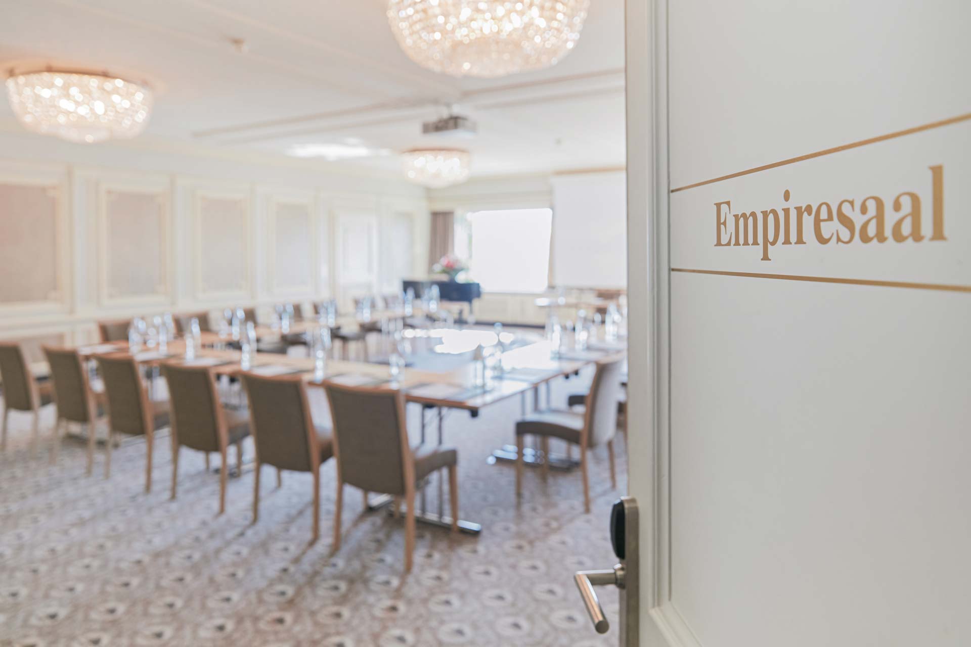 Porte ouverte avec inscription « Salle Empire », vue sur une grande pièce avec moquette à motifs, motifs ronds en cristal, boiseries blanches et grandes fenêtres panoramiques, tables et chaises au centre.