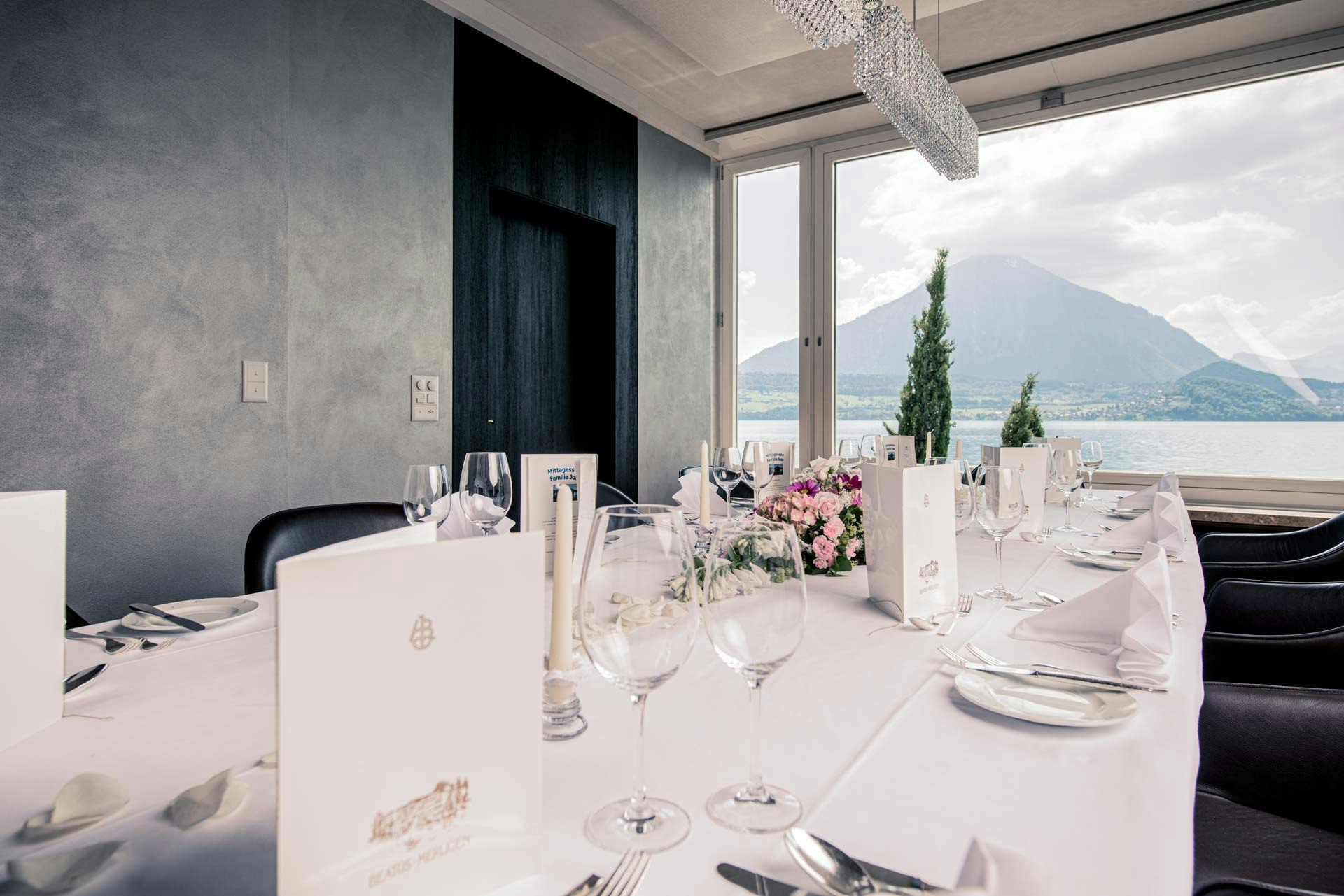 Salle aux murs sombres, au centre une grande table couverte d'une nappe blanche, grande fenêtre avec vue sur le lac