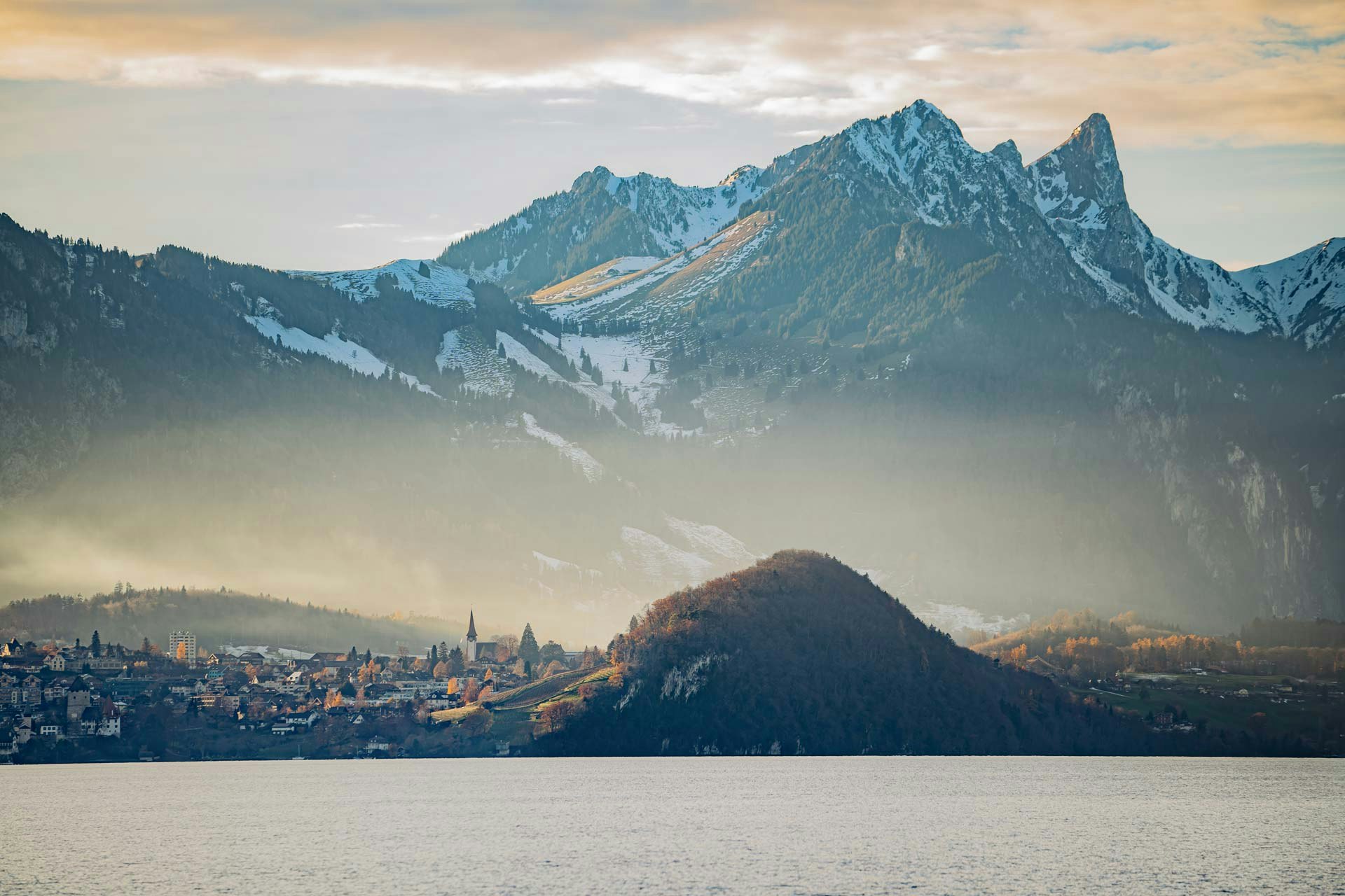 Mächtige Berge mit Schnee im Hintergrund, davor ein Dorf und ein bewaldeter Hügel, ganz vorne der Thunersee