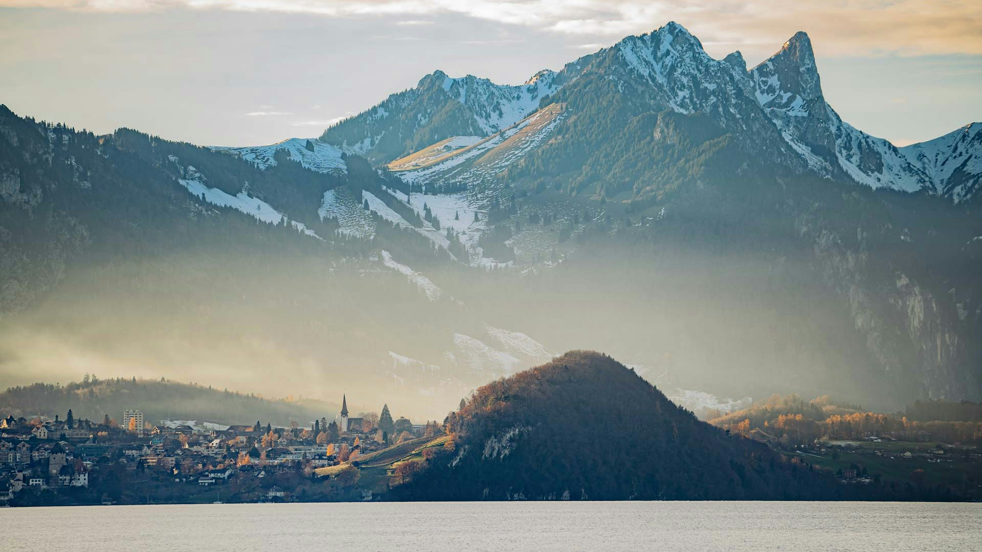 Blick auf See und Berge Mächtige Berge mit Schnee im Hintergrund, davor ein Dorf und ein bewaldeter Hügel, ganz vorne der Thunersee