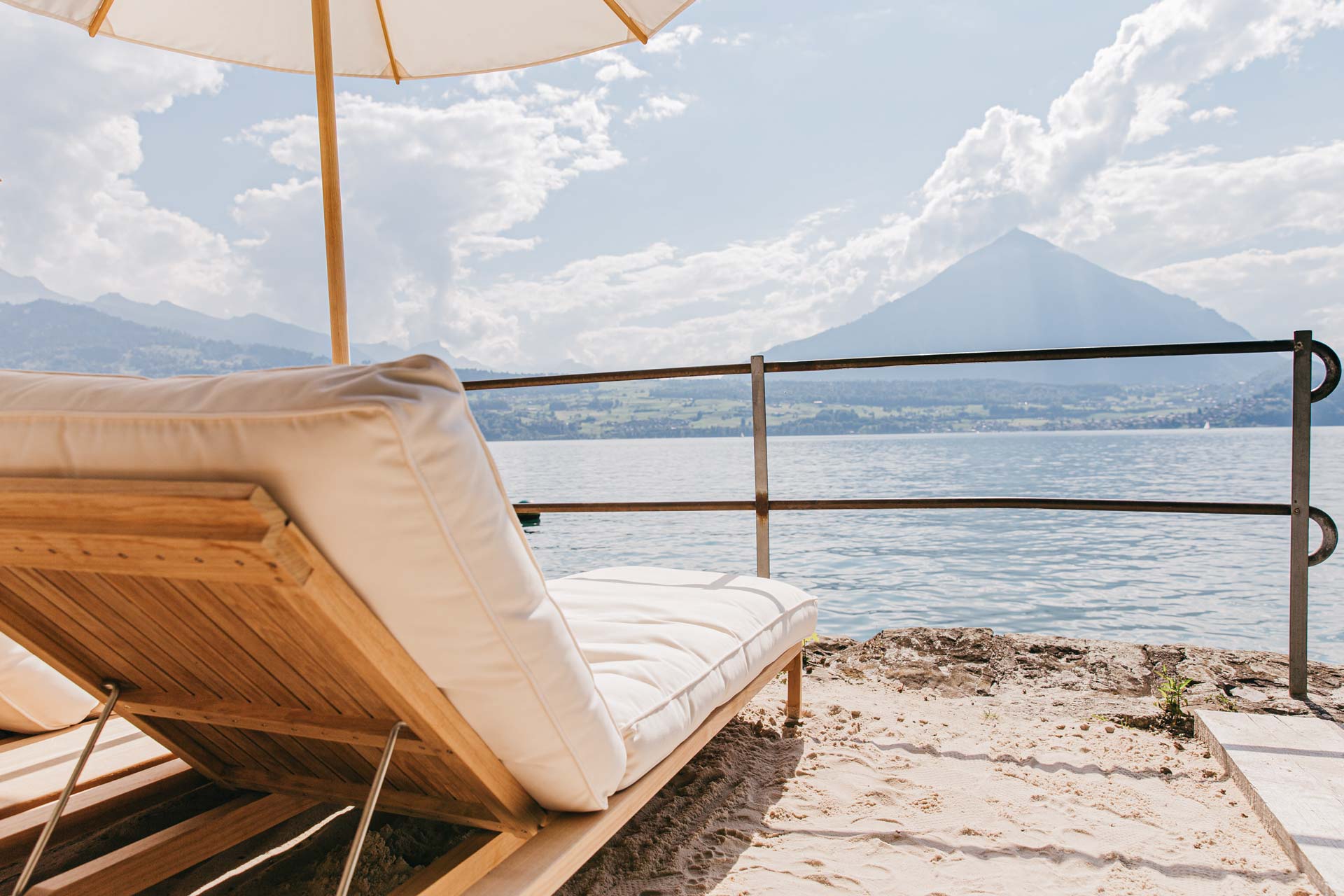 Sandstrand und gepolsterte weisse Liege mit Sonnenschirm, Blick auf See und Berg