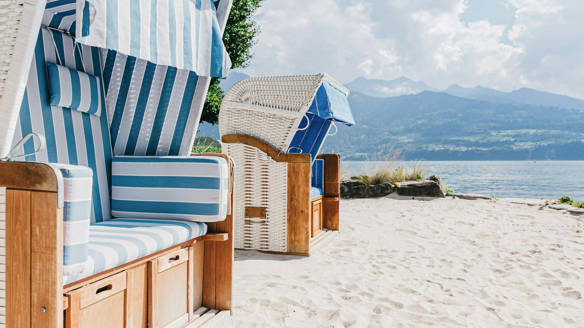 Beach chairs on the hotel beach at the Hotel BEATUS Two blue and white striped beach chairs on the left, sandy beach in front, lake and mountains in the background