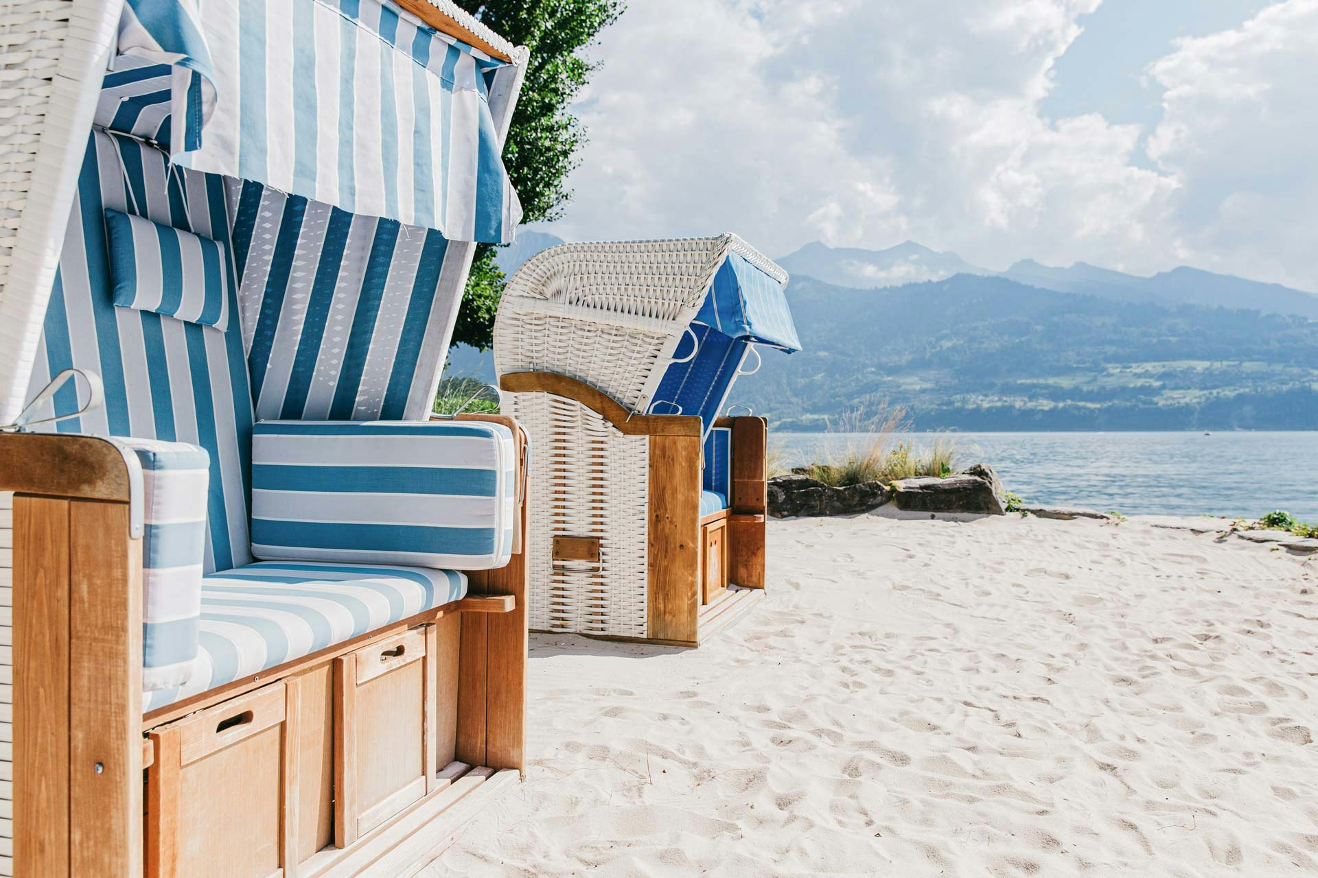 Links zwei blau-weiss gestreifte Strandkörbe, davor Sandstrand, im Bildhintergrund See und Berge