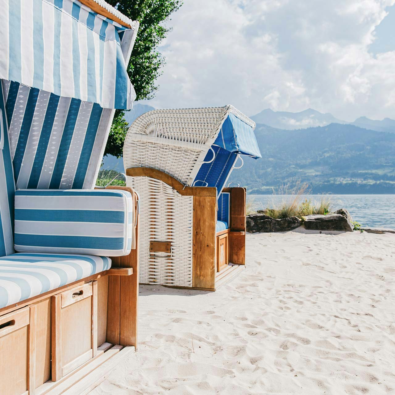 Strandkörbe am Hausstrand des Hotel BEATUS Links zwei blau-weiss gestreifte Strandkörbe, davor Sandstrand, im Bildhintergrund See und Berge