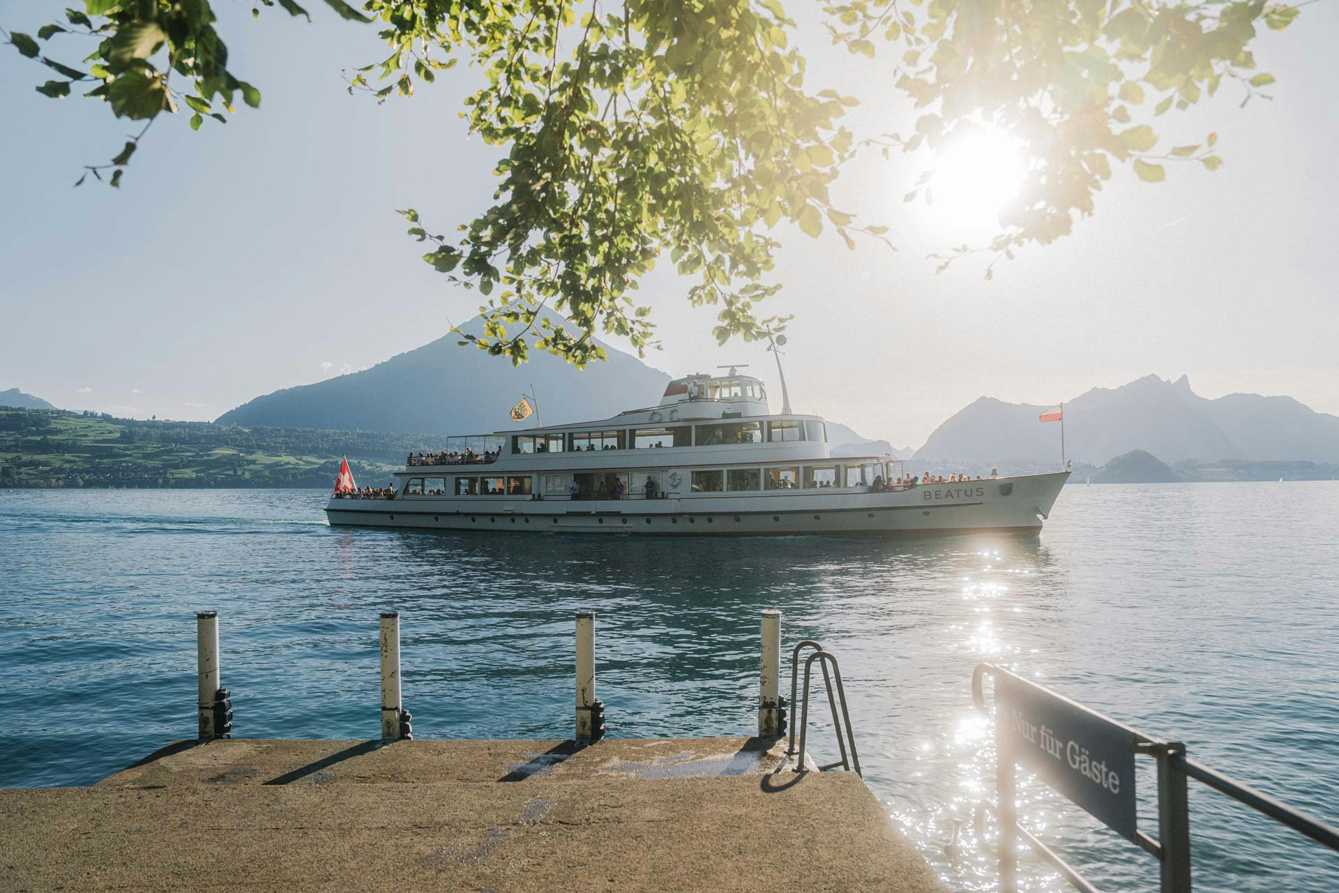 Vorne der Steg nur für Hotelgäste, dahinter im Sonnenlicht glitzernd der See und ein Ausflugsschiff