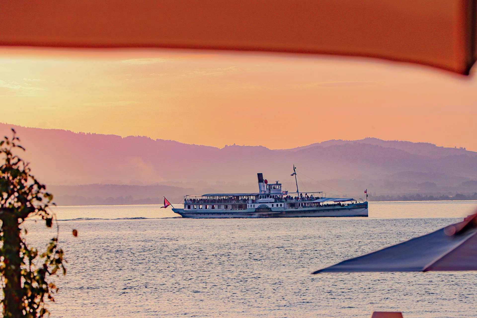 Ausflugsschiff am See, dahinter die Berge, darüber ein Teil eines Sonnenschirmes, alles in gelb-oranges Licht getaucht