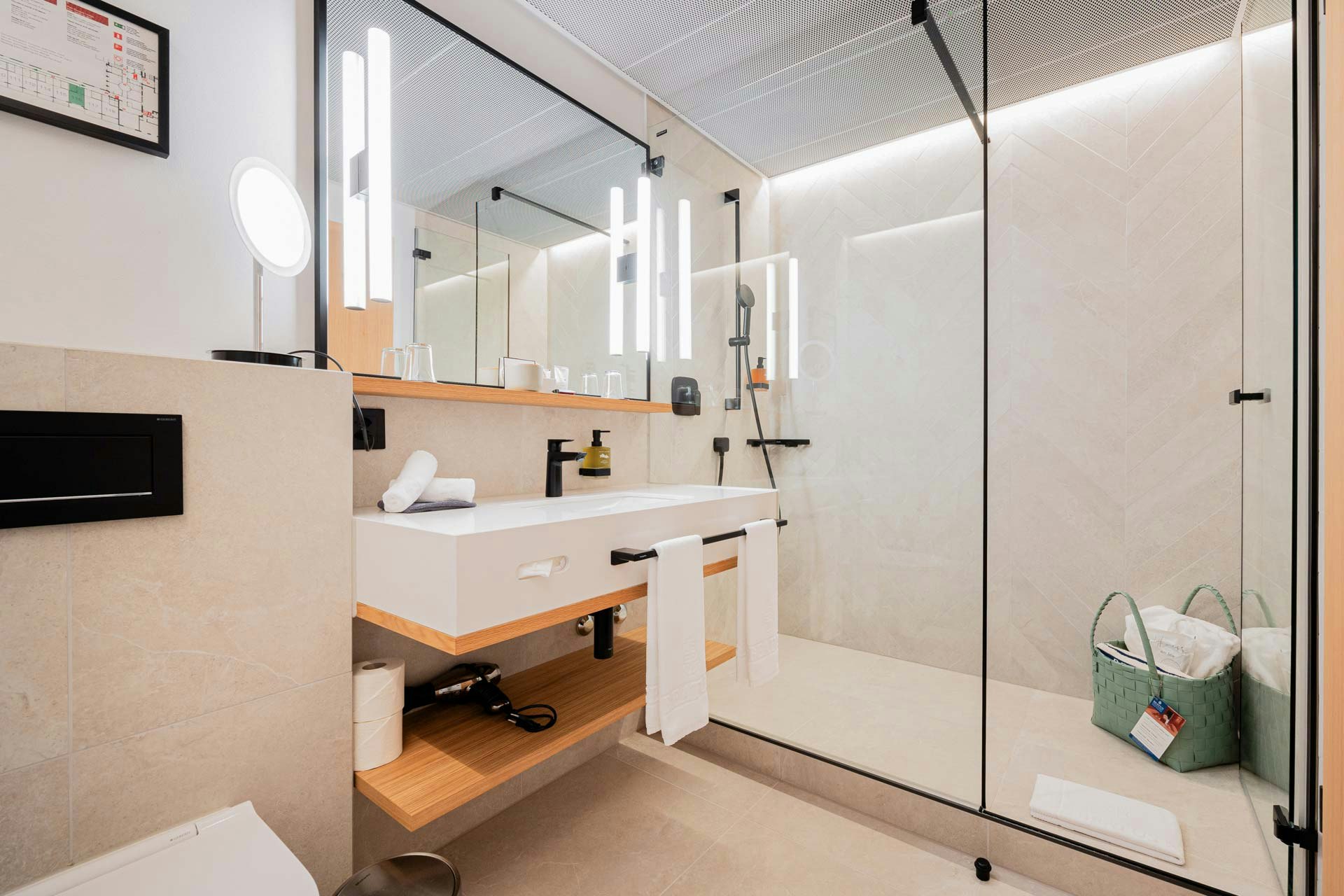 Hell gefliestes Badezimmer mit zwei Waschbecken und Ablage auf hellem Holz, grosser Spiegel, Dusche mit Glaswand, darin eine Badetasche