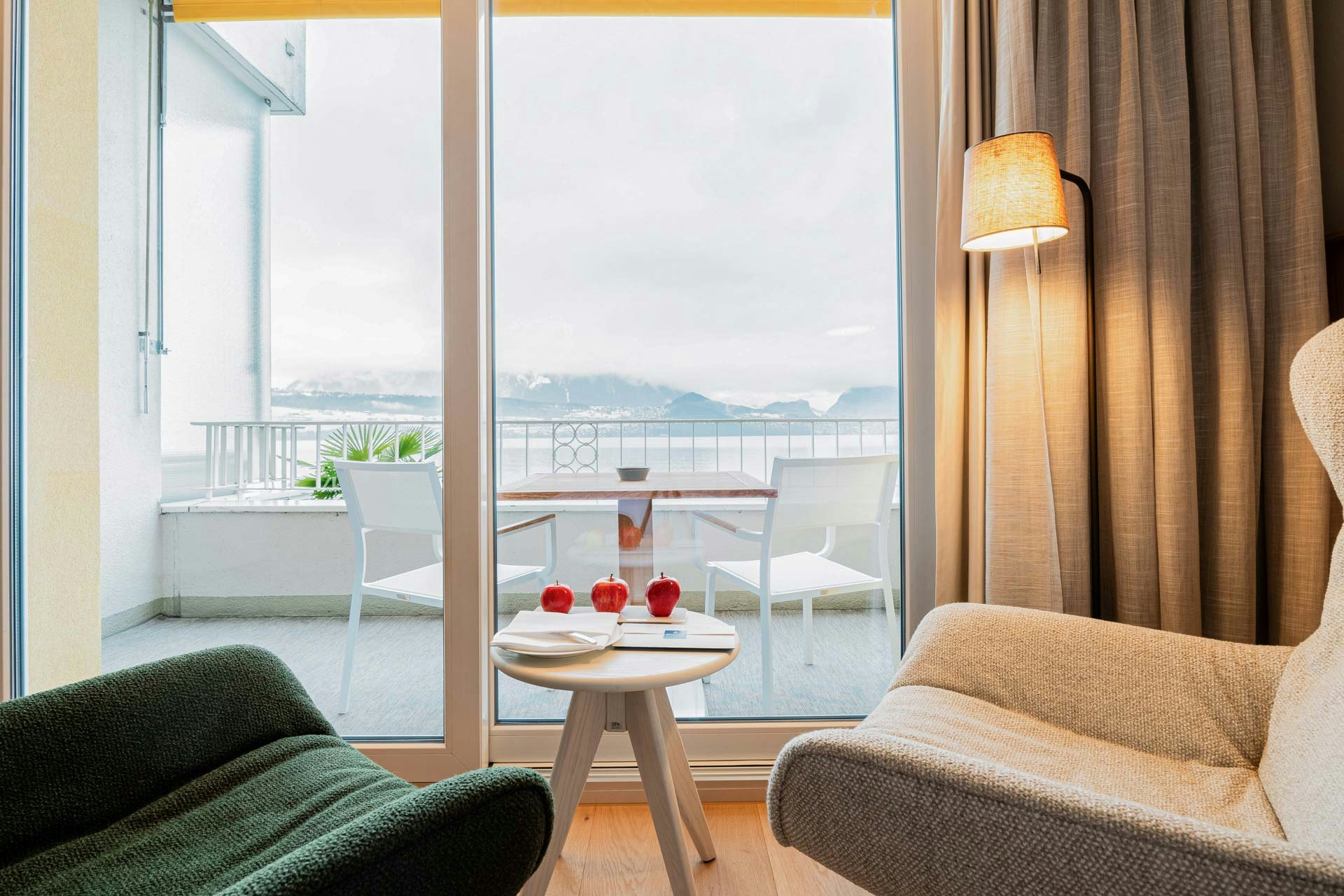 Vue sur deux fauteuils à oreilles, entre eux une petite table en bois blanc avec trois pommes rouges, à l'arrière une fenêtre panoramique sur le balcon