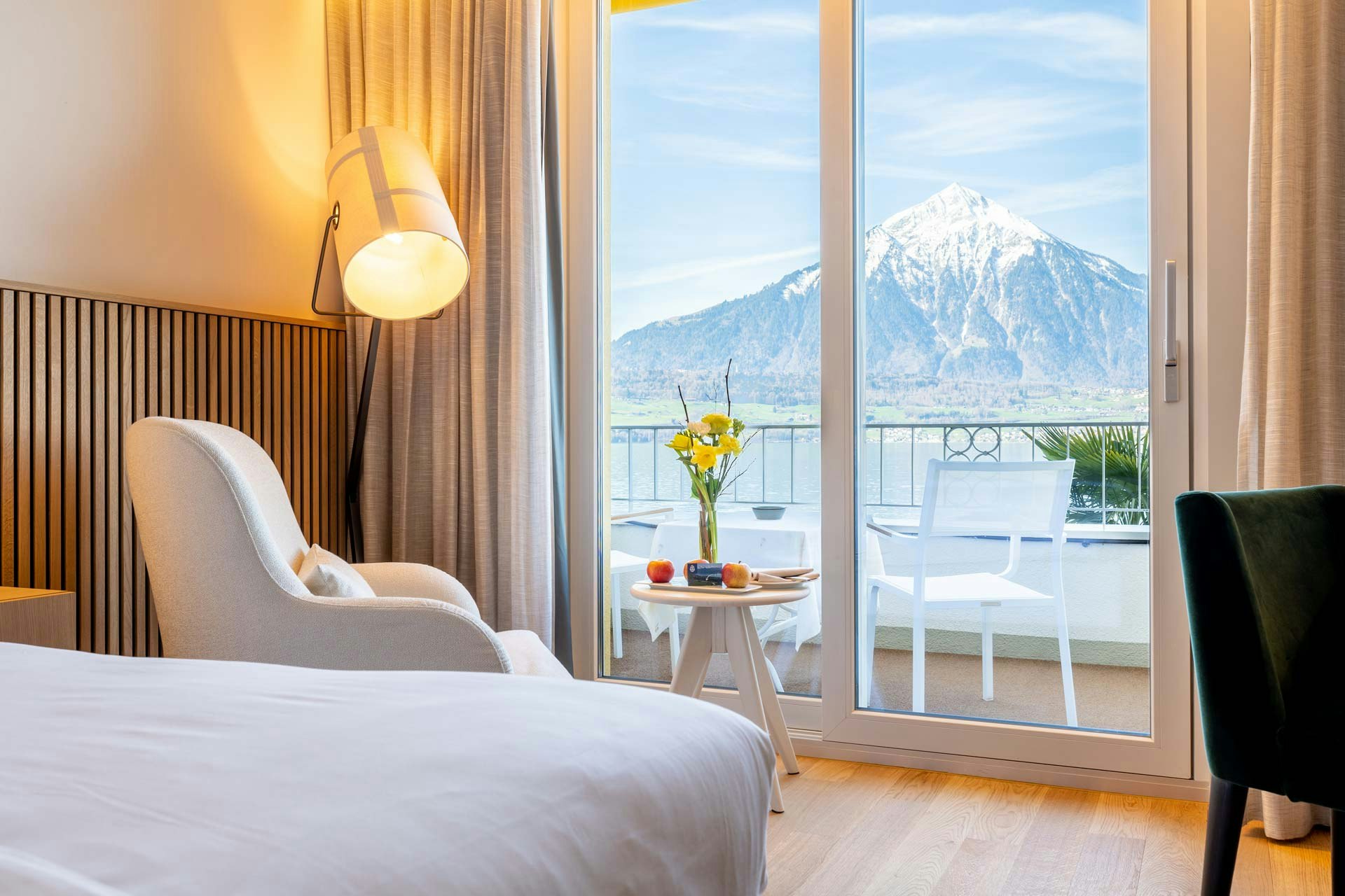 Eine Stehlampe beleuchtet einen Ohrensessel und einen runden Holztisch mit Äpfeln und Blumen, grosse Balkontür mit Blick auf den See und einen Berg