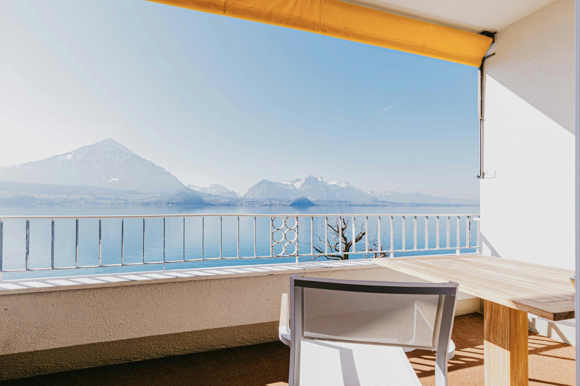 Balcony with a view of the blue lake and the mountains, above a piece of a yellow sun blind, on the right a wooden table and a chair