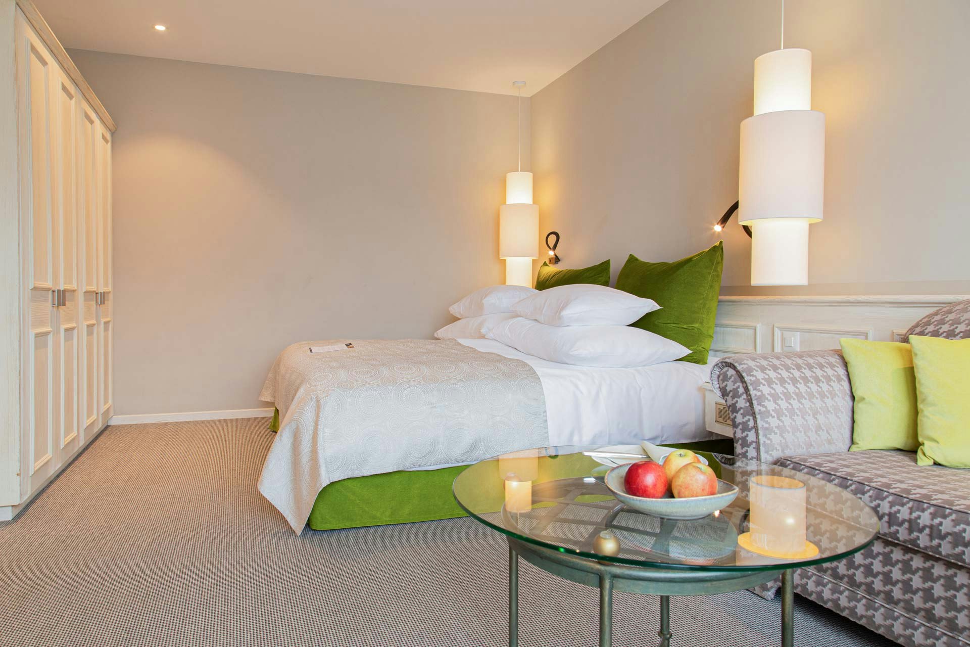 Chambre avec moquette et armoire murale blanche, à droite au premier plan une table ronde en verre et un canapé, au fond un lit double