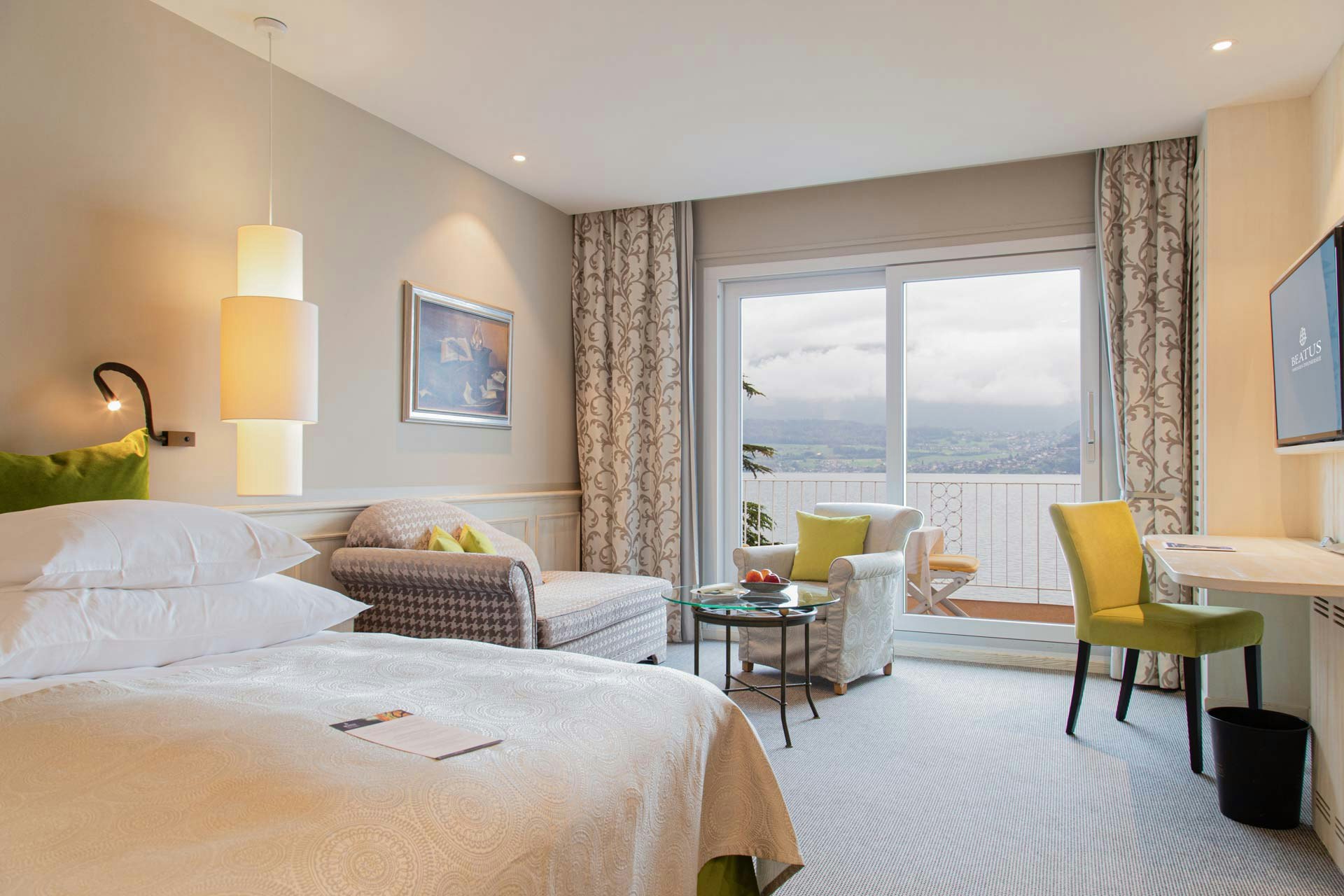 Chambre avec lit double, lampe suspendue, derrière canapé, fauteuil rembourré, table en verre, à droite contre le mur un écran plat, en dessous une chaise devant une table pliante, derrière fenêtre panoramique avec vue sur le lac,