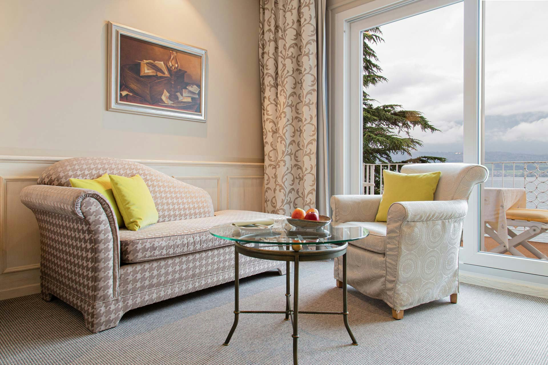 Chambre avec tapis, canapé, fauteuil rembourré et table en verre, une nature morte au mur, fenêtre panoramique avec vue sur un balcon