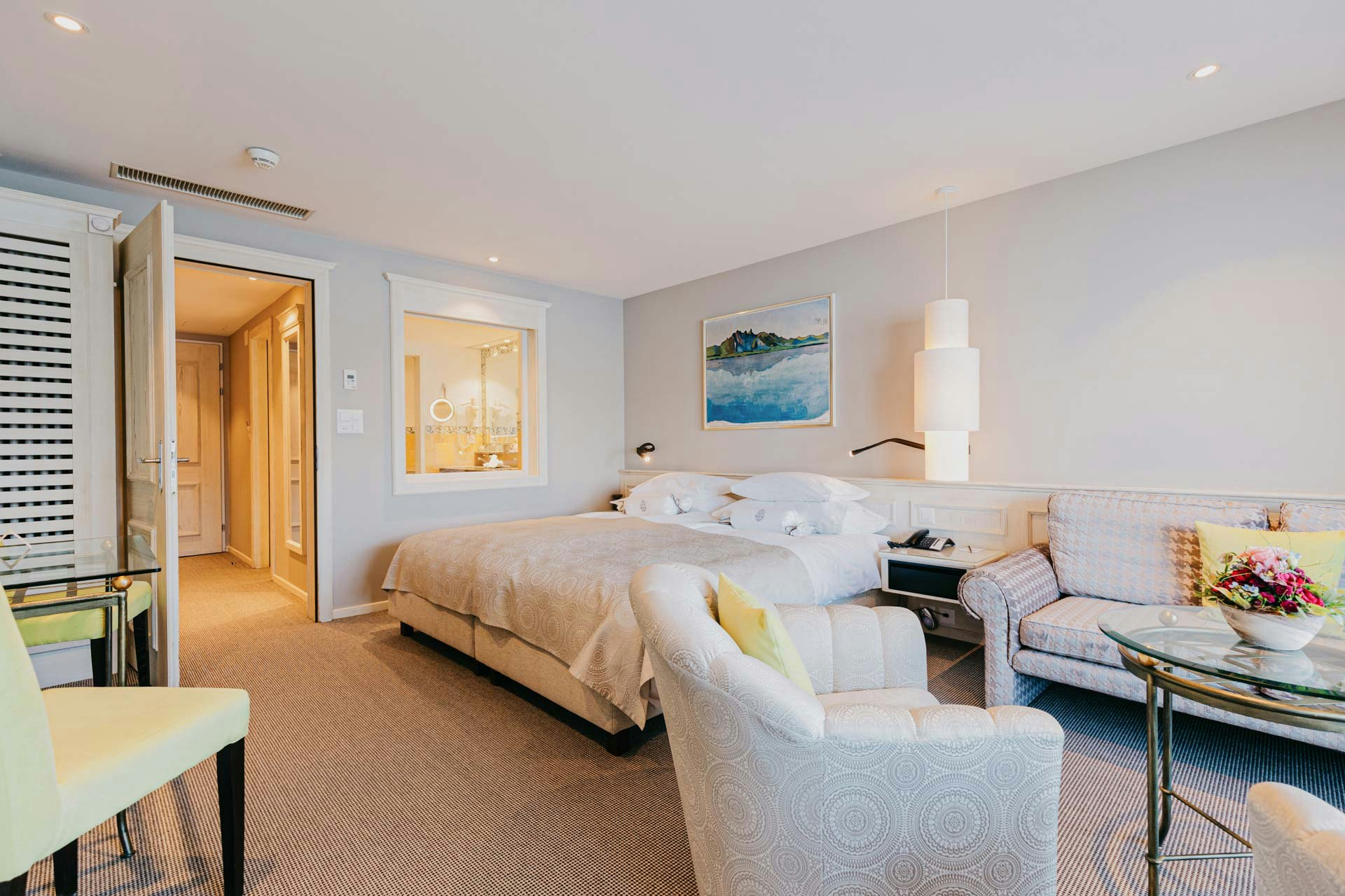Une chambre avec moquette, au premier plan une table en verre et un meuble rembourré, derrière un lit double avec une lampe suspendue et une peinture de paysage, au fond une porte et une baie vitrée donnant sur une autre pièce.