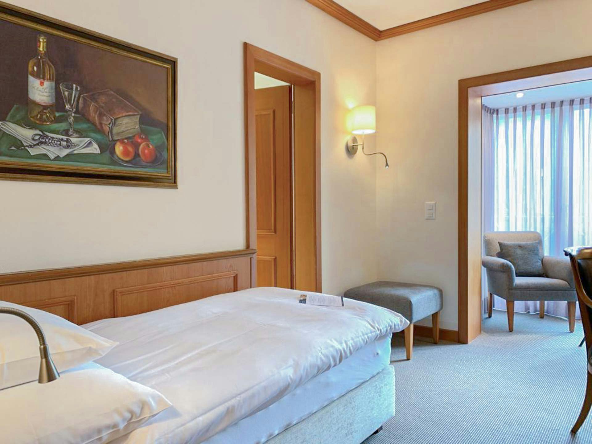 Zimmer mit Teppichboden, Einzelbett, darüber ein Stillleben, Blick in einen Nebenraum mit Polstersessel und Fenster