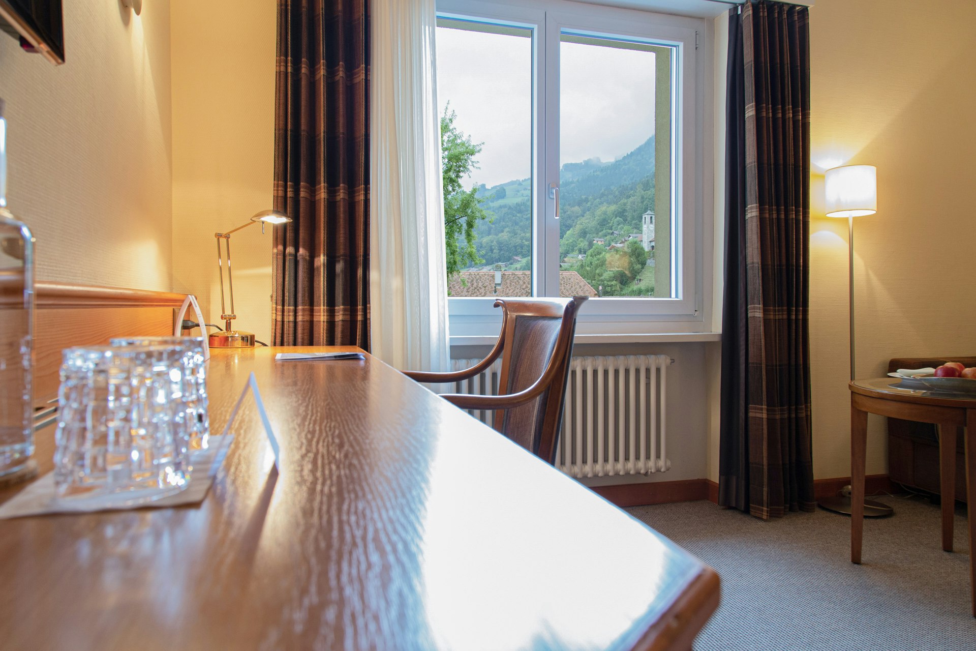 Zimmer mit Teppichboden, Schreibtisch aus Holz, davor ein Stuhl, hinten ein Fenster mit Vorhängen