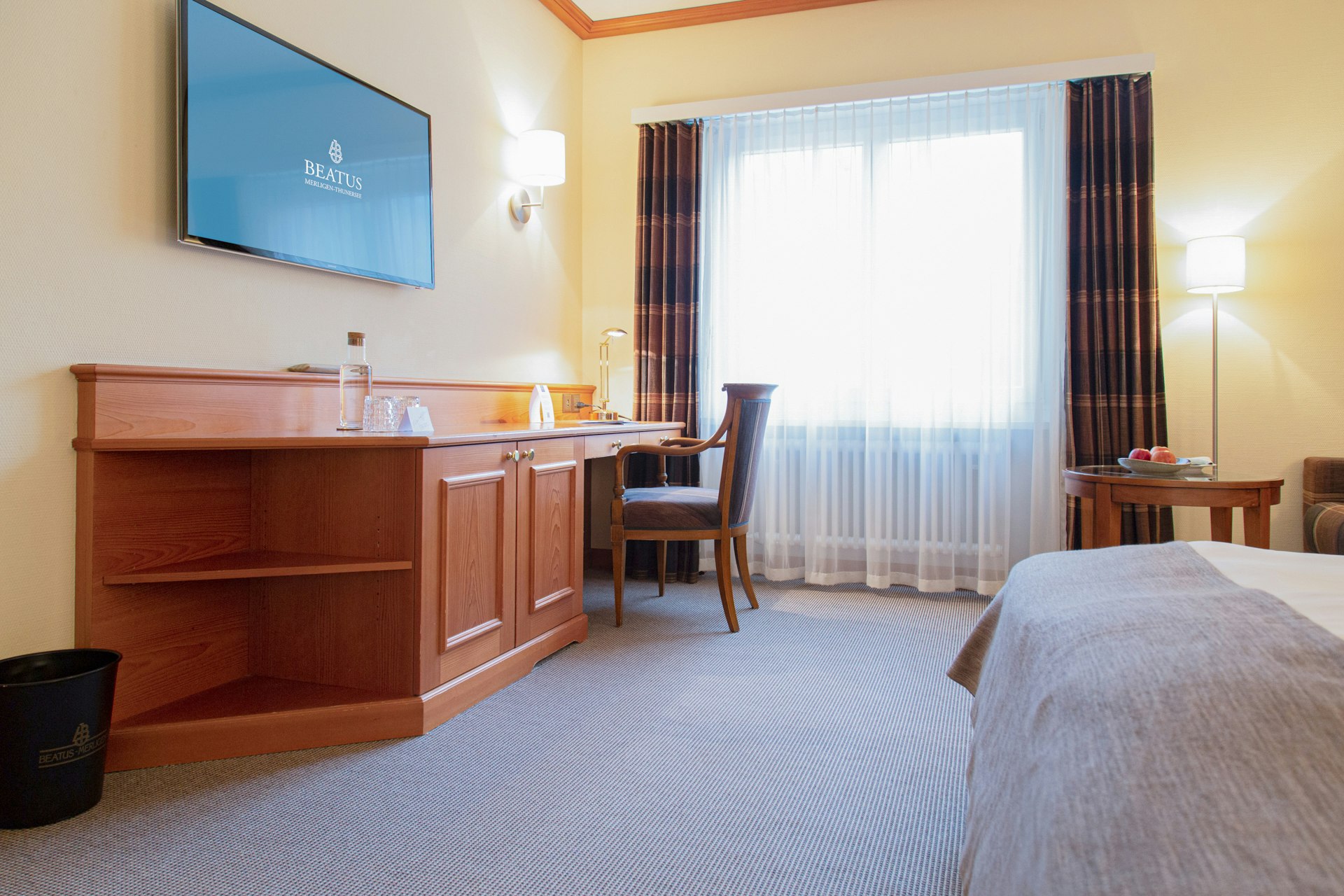 Zimmer mit Teppichboden, Schreibtisch aus Holz, davor ein Stuhl, darüber ein Flachbildschirm, hinten ein Fenster mit Vorhängen, rechts angeschnitten ein Bett