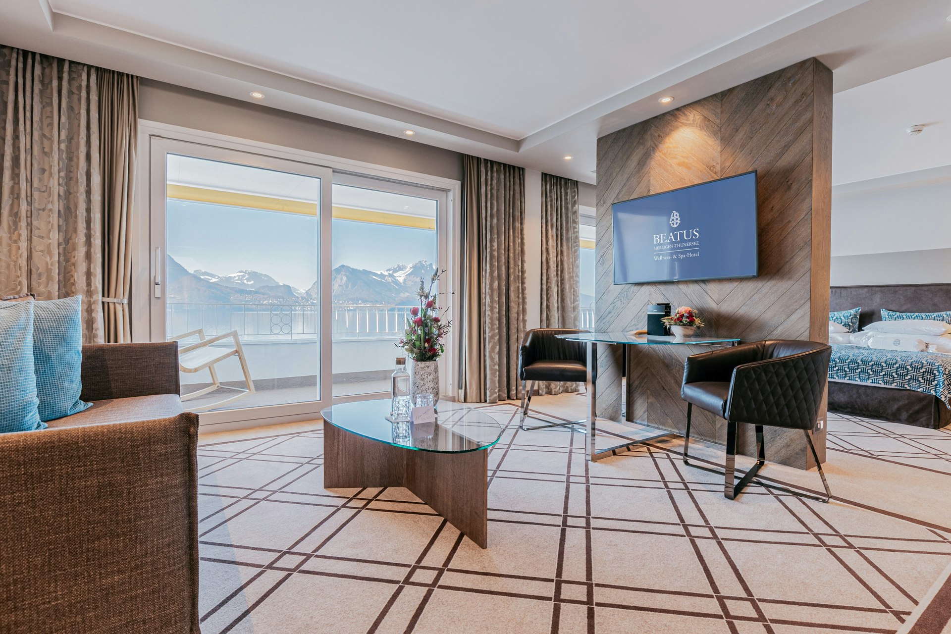 Zimmer mit Teppich, Glastisch, Blick auf den Balkon und Berge, vor einer Trennwand ein kleiner Tisch mit bequemen Stuhl, darüber angebracht ein Flachbildschirm, dahinter ein Bett